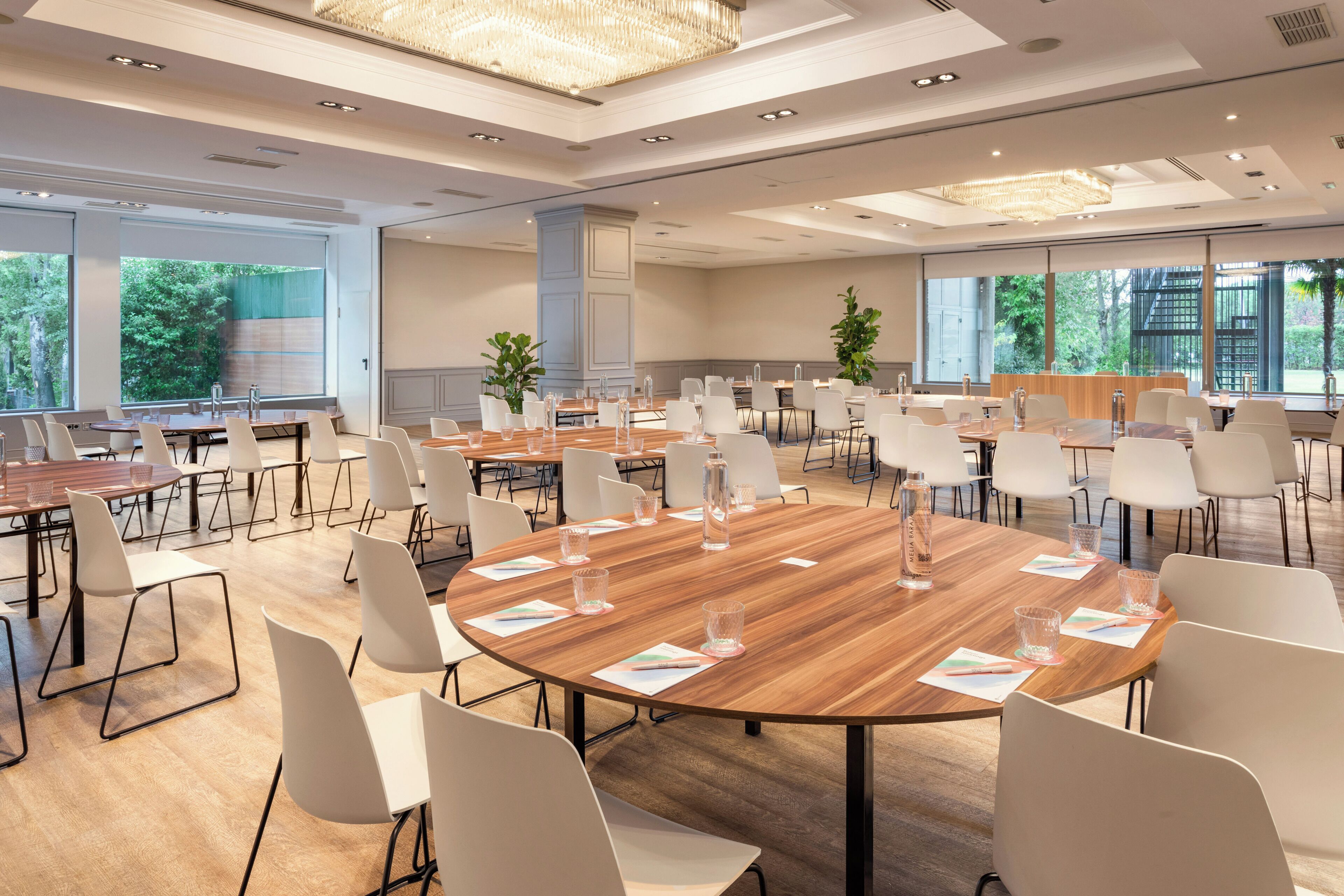 Ein geräumiger Konferenzraum mit mehreren Tischen, weißen Stühlen und großen Fenstern mit Blick auf Grünflächen.