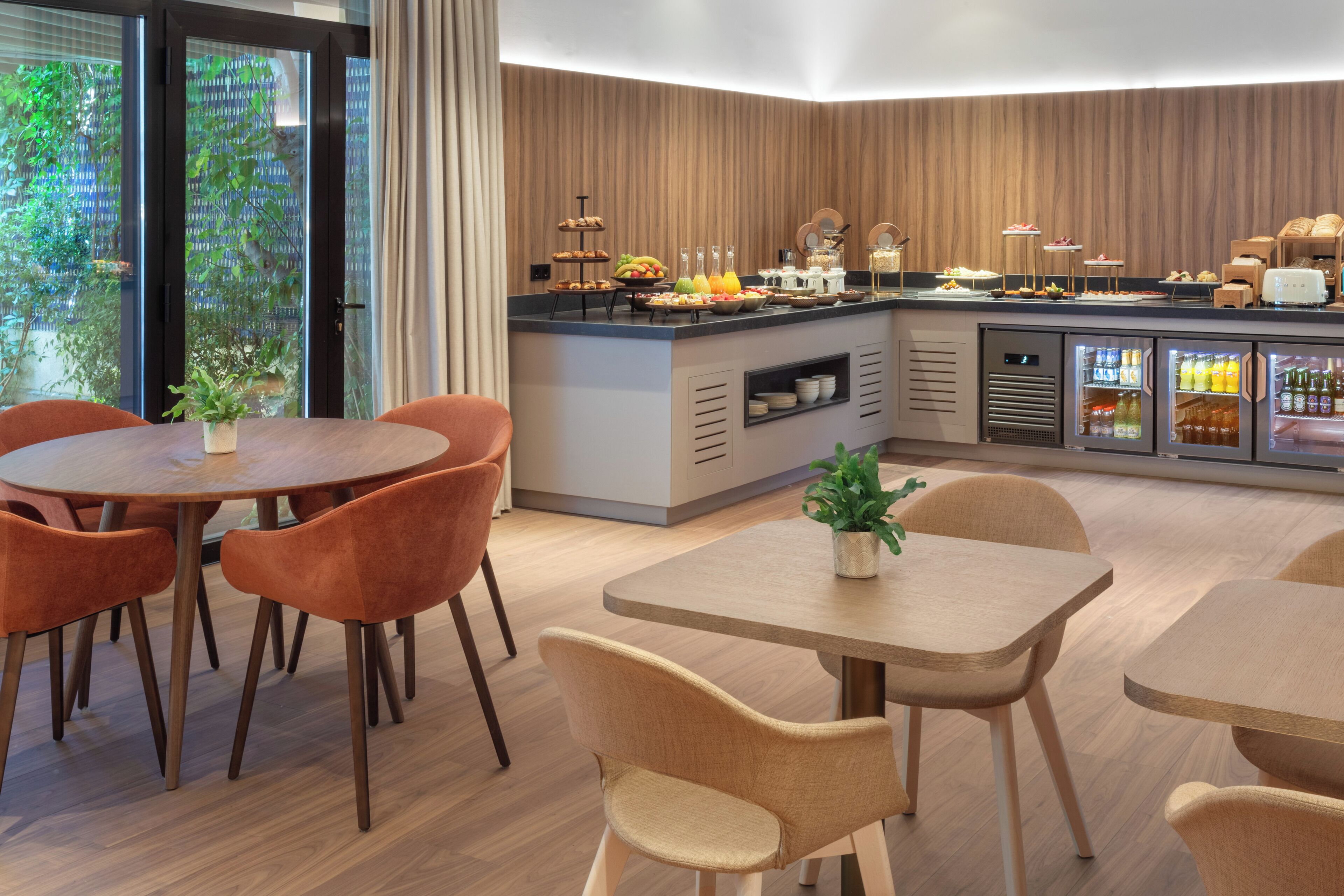 Ein zeitgemäßer Essbereich mit Holztischen und -stühlen, eine Selbstbedienungsbar mit Speisen und ein mit Getränken gefüllter Kühlschrank.