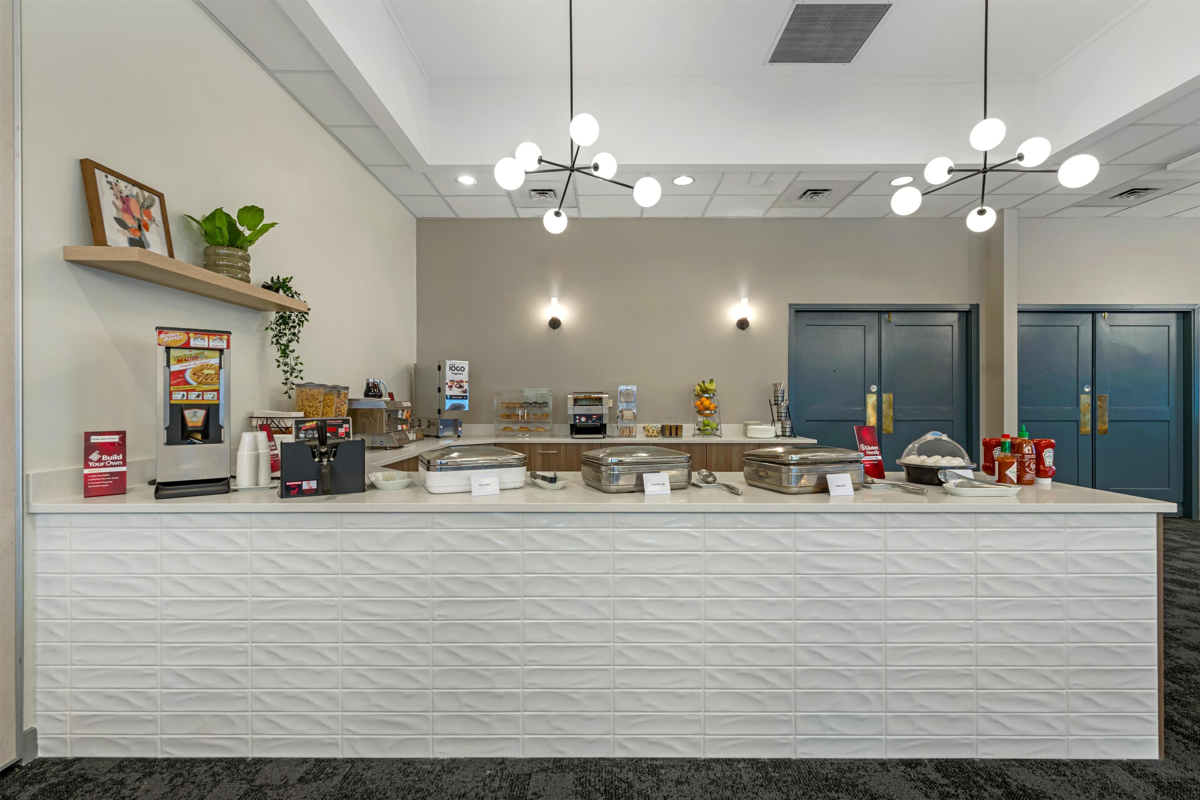 A hotel reception area with a white reception desk, multiple light fixtures, a coffee machine, and a plant on a shelf.
