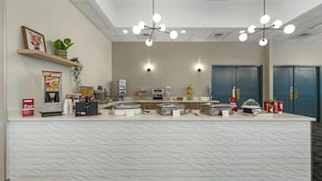 A hotel reception area with a white reception desk, multiple light fixtures, a coffee machine, and a plant on a shelf.