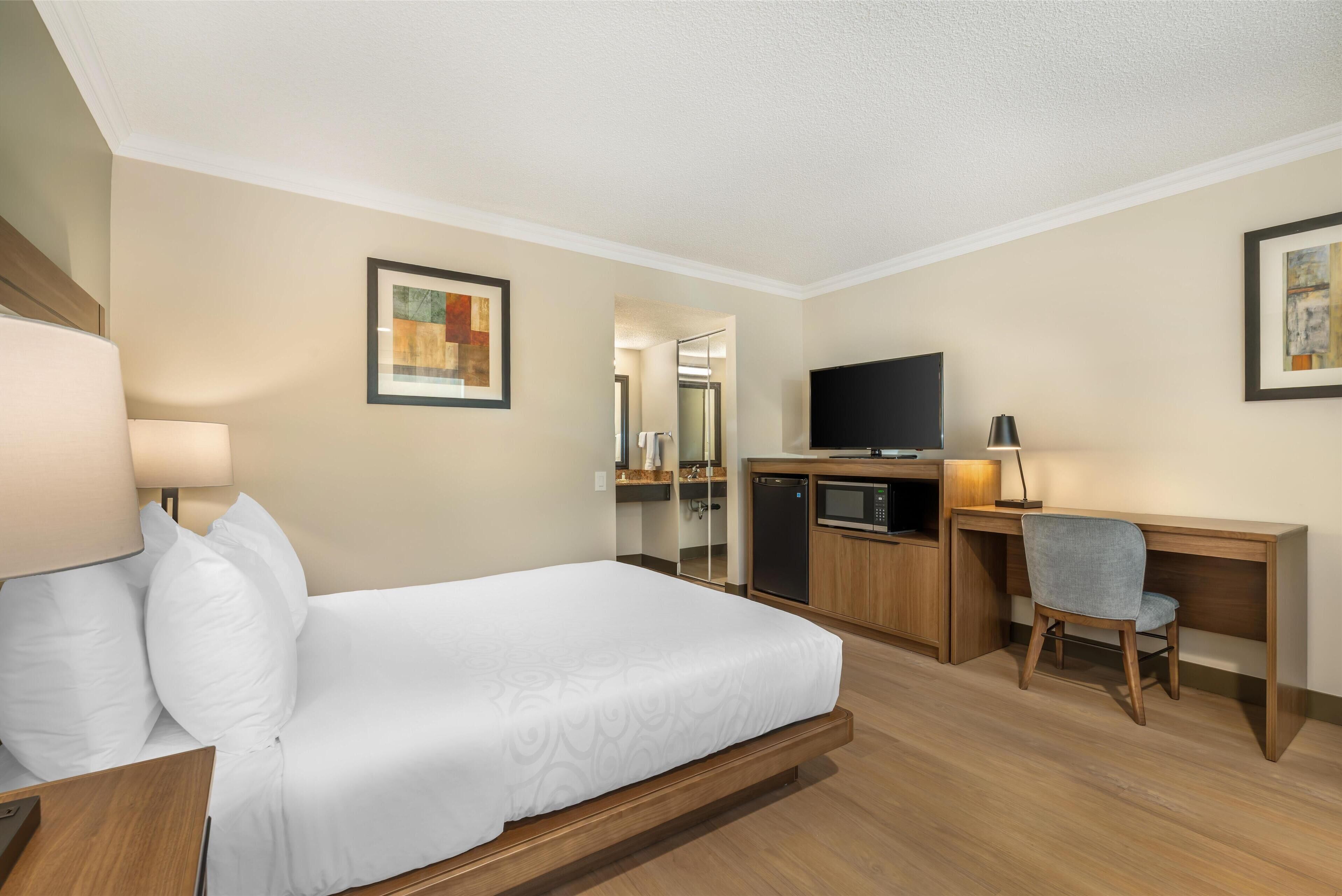 A hotel room with a bed, desk, chair, TV, and a small kitchenette.