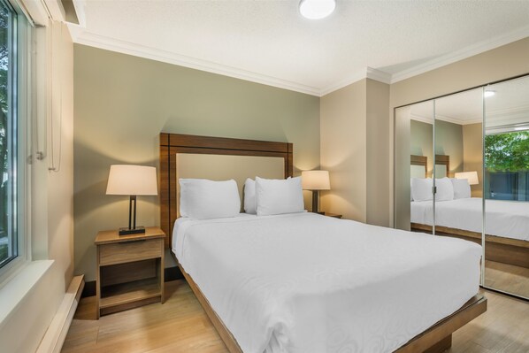 A neatly made bed with white linens, two bedside tables with lamps, a large mirror, and a window with a view of greenery.