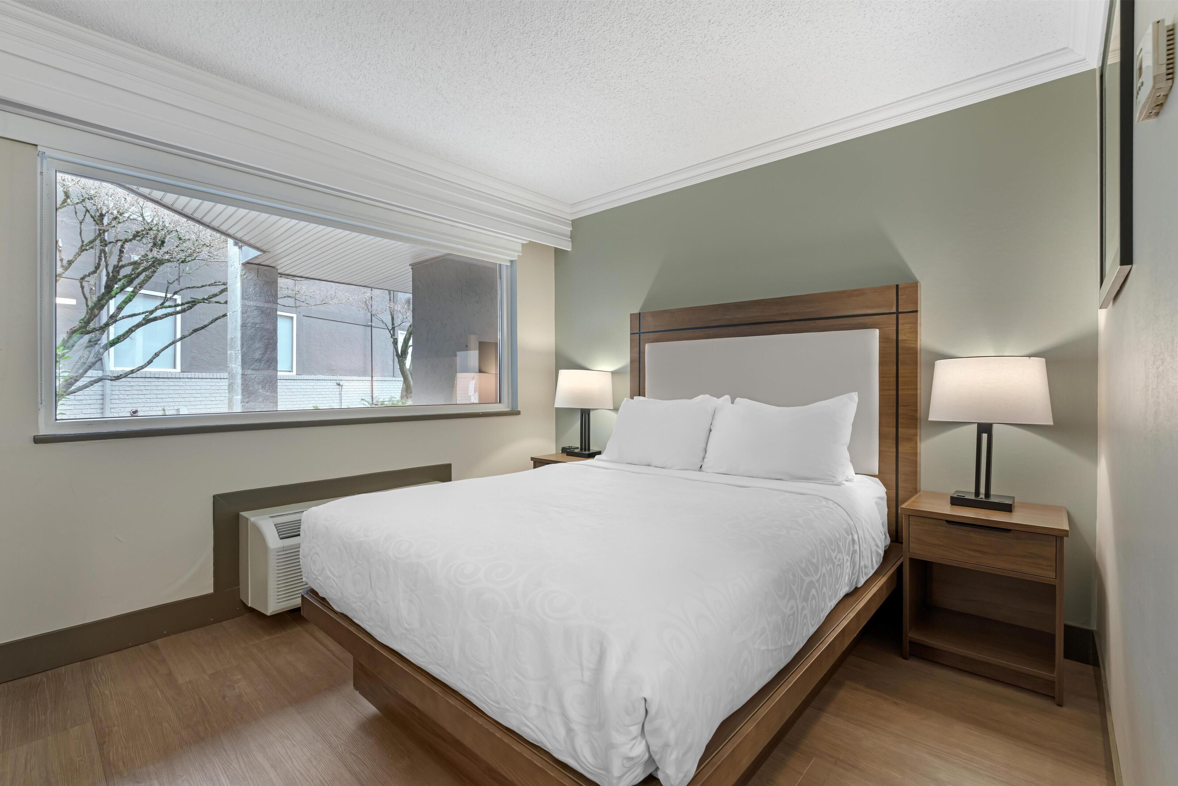 A modern hotel room with a large bed, two bedside tables with lamps, a window with a view of the outside, and a built-in heater.