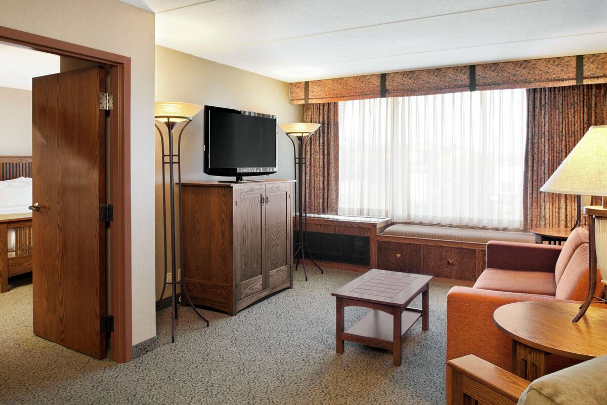 A hotel room with a wooden door, a television on a cabinet, a sofa, a small table, and a window with curtains.