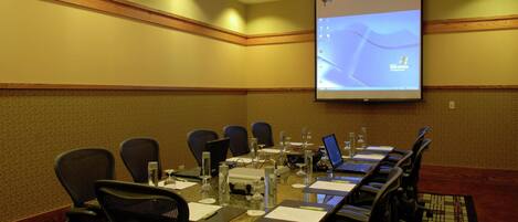 Un salón de conferencias con una mesa larga, sillas, una pantalla de proyector y una computadora portátil.