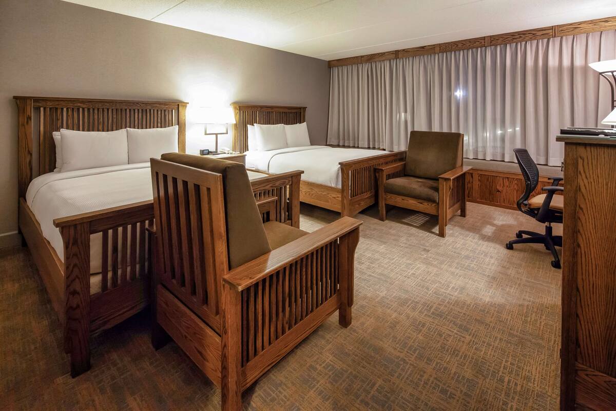 A hotel room with two wooden beds, a wooden desk, a chair, and a small table.