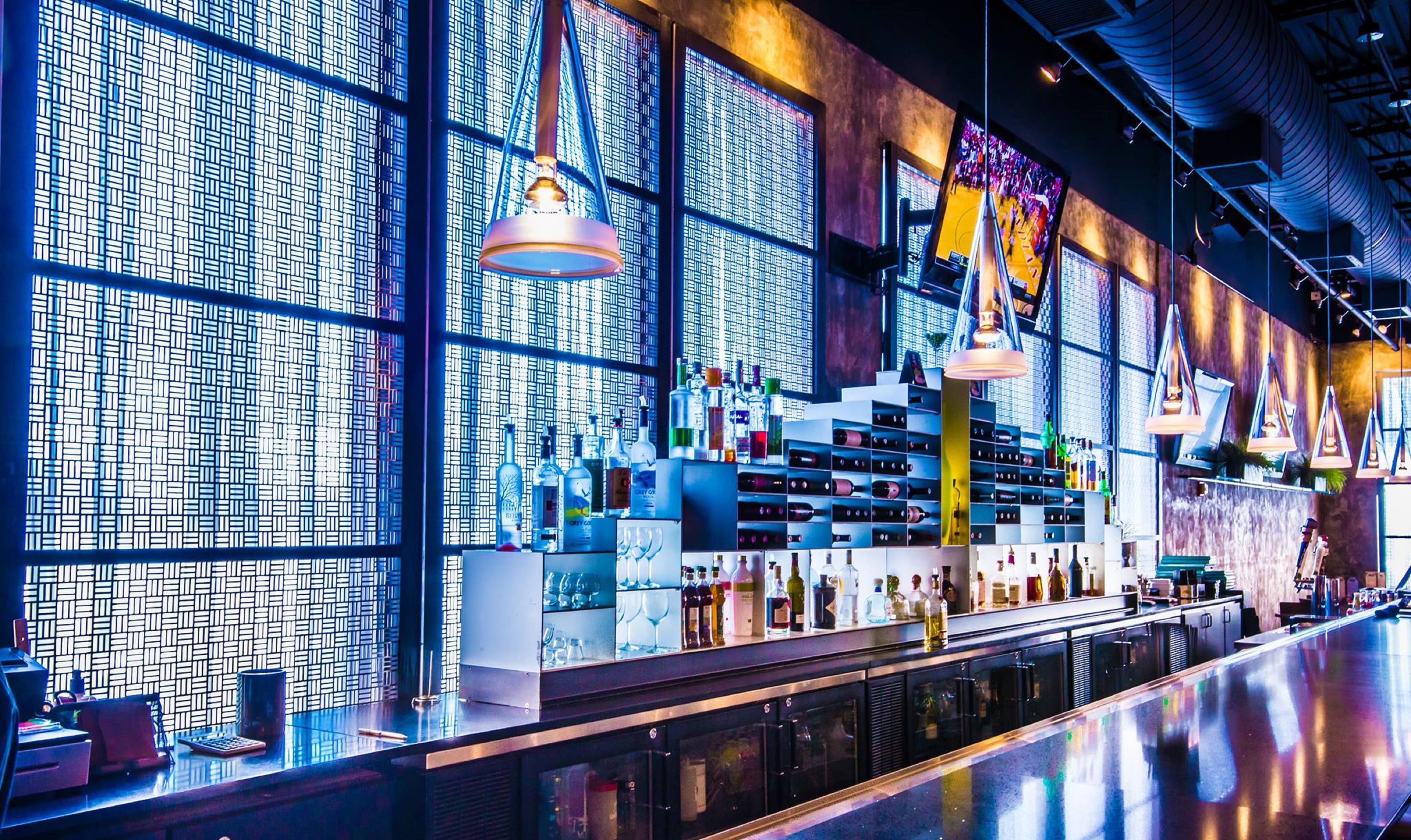 A bar with a patterned wall, shelves of bottles, and hanging pendant lights.