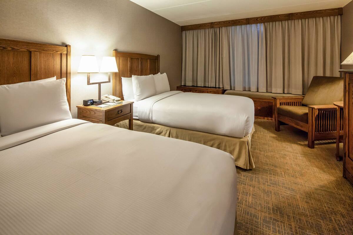 A hotel room with two beds, a nightstand with a lamp, a chair, and a large window with curtains.
