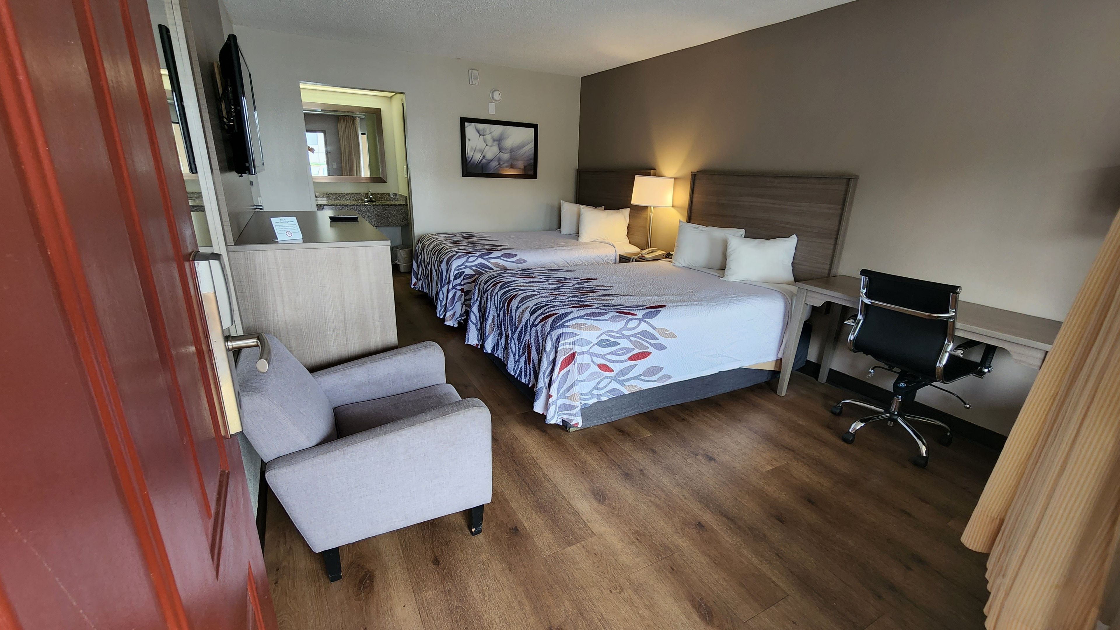 A hotel room with a bed, two bedside tables, a desk with a chair, a TV, and a bathroom visible through an open door.