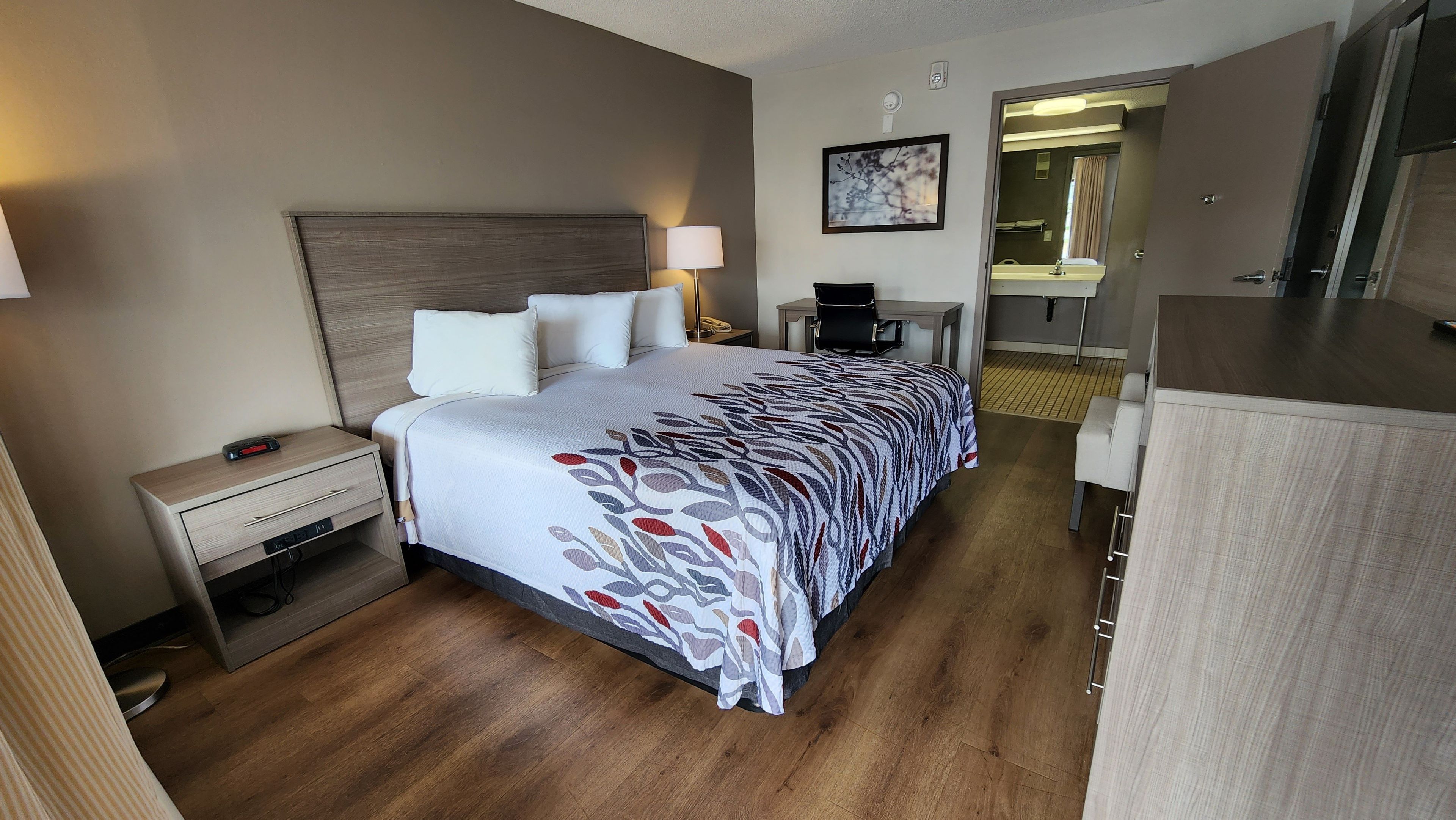 A hotel room with a bed, bedside table, lamp, and a small desk with a chair.