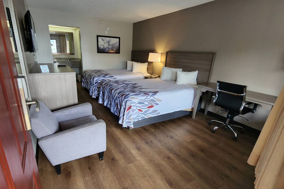 Quarto de hotel com cama de casal, escrivaninha, cadeira, televisão e banheiro visível por uma porta aberta.