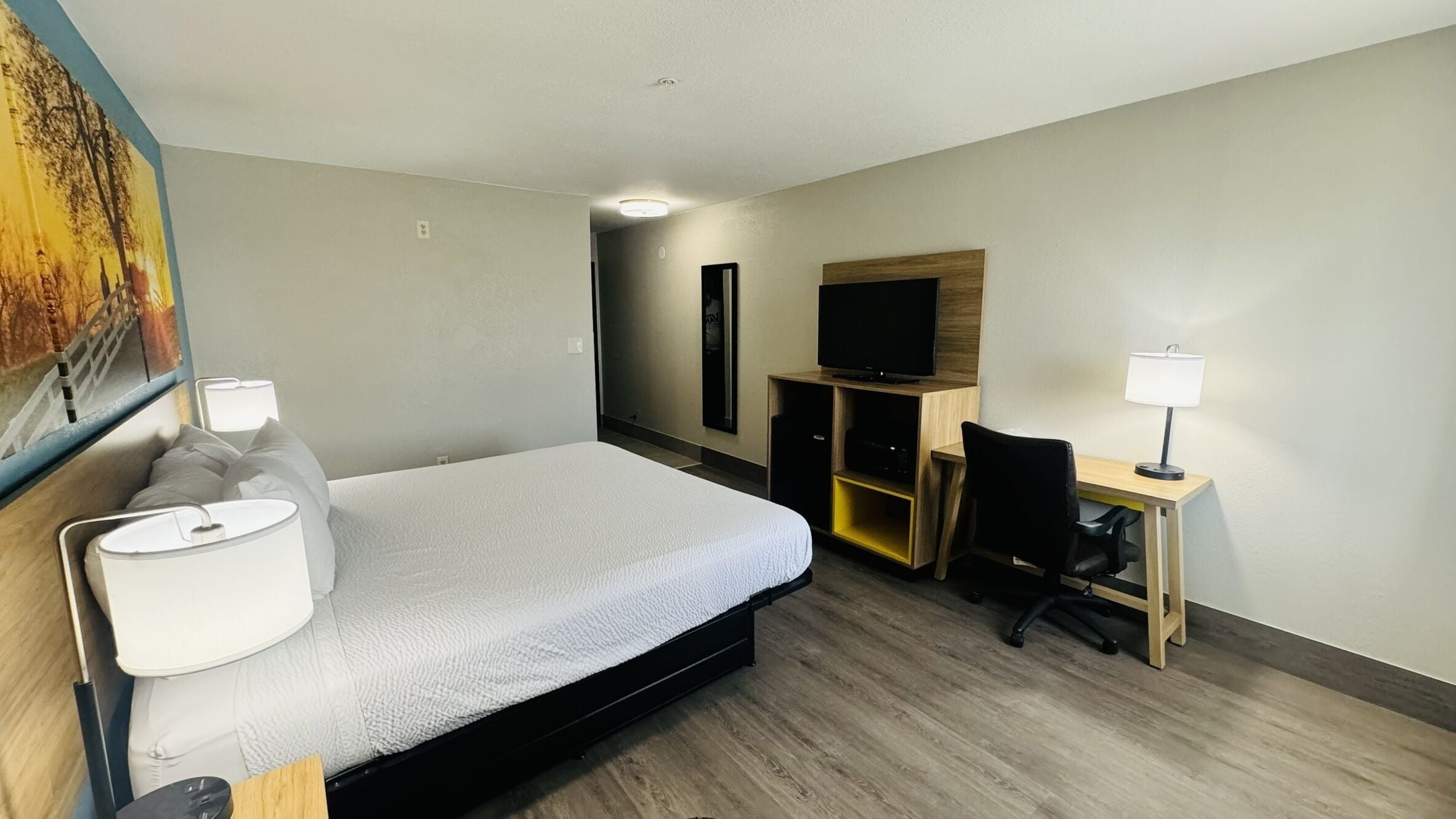 A hotel room with a bed, a desk with a chair, a TV, and a painting on the wall.