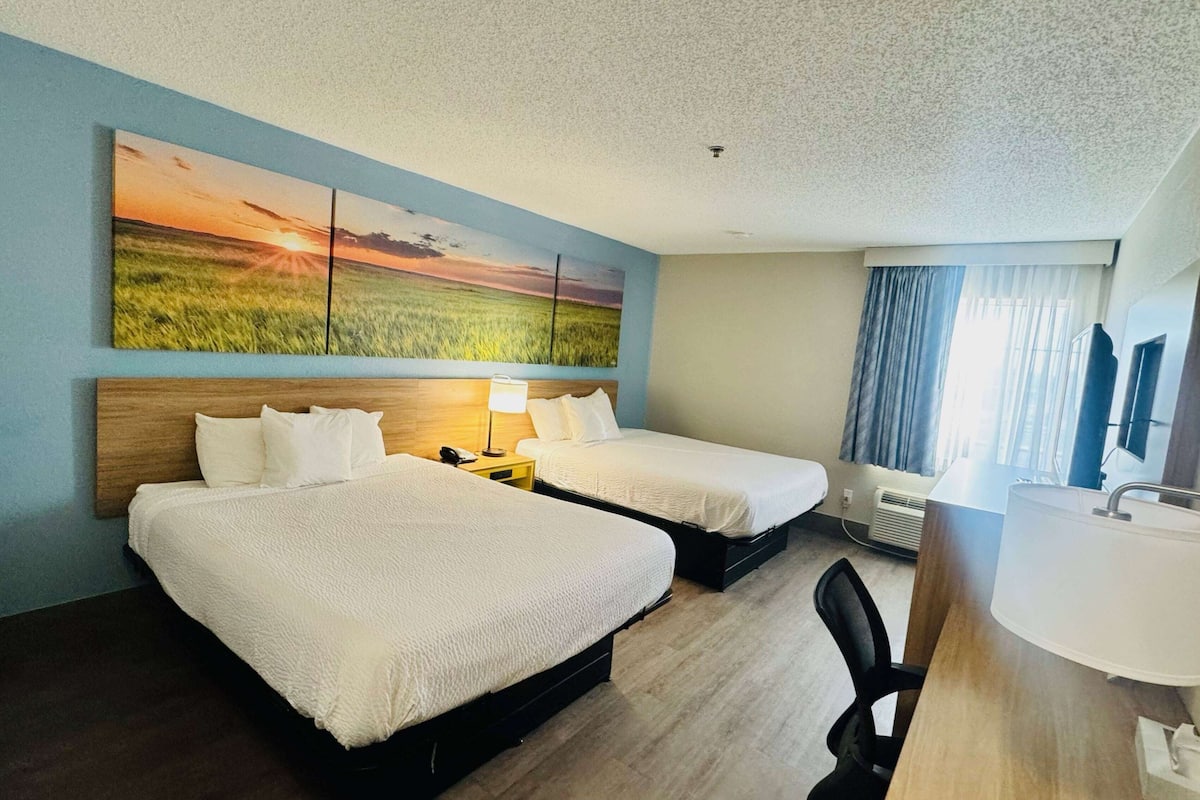 A hotel room with two beds, a desk, a chair, a lamp, and a large painting on the wall.
