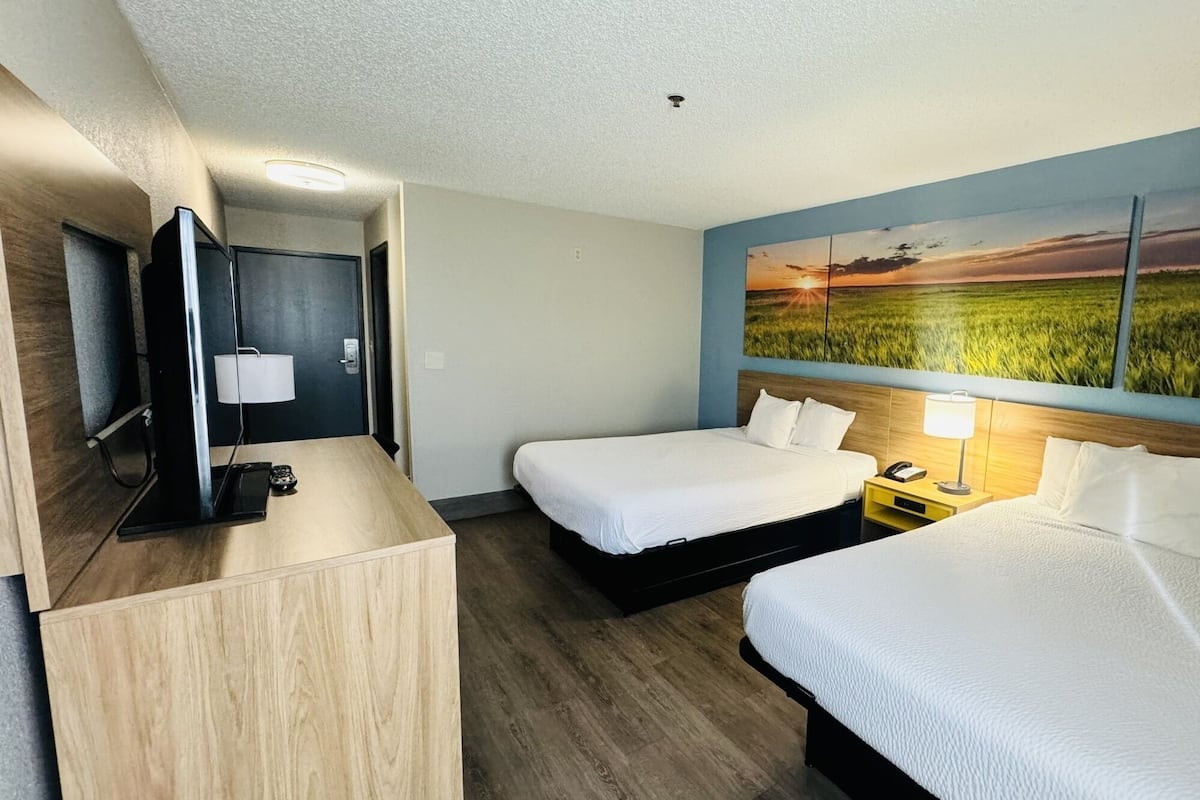 A hotel room with two beds, a TV, a desk, and a painting of a sunset over a field.