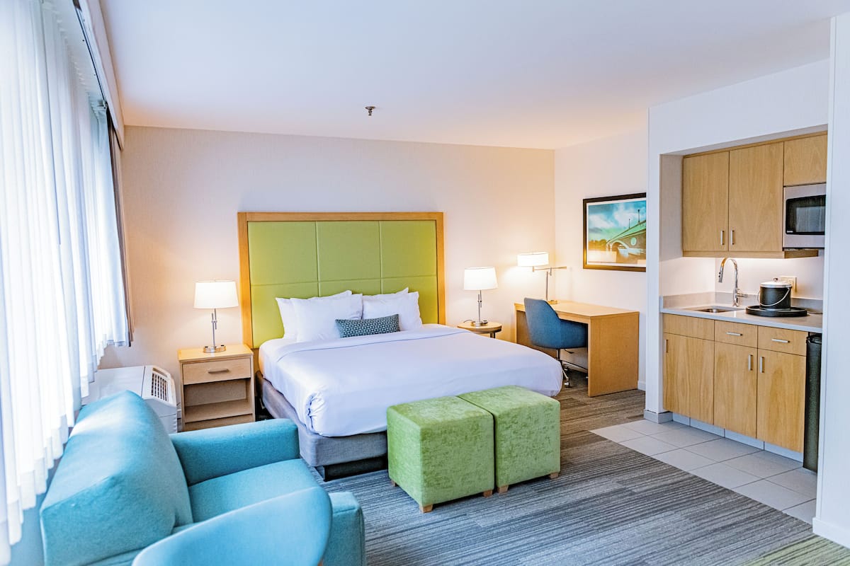 A hotel room with a large bed, a desk, a chair, a kitchenette, and a window with curtains.