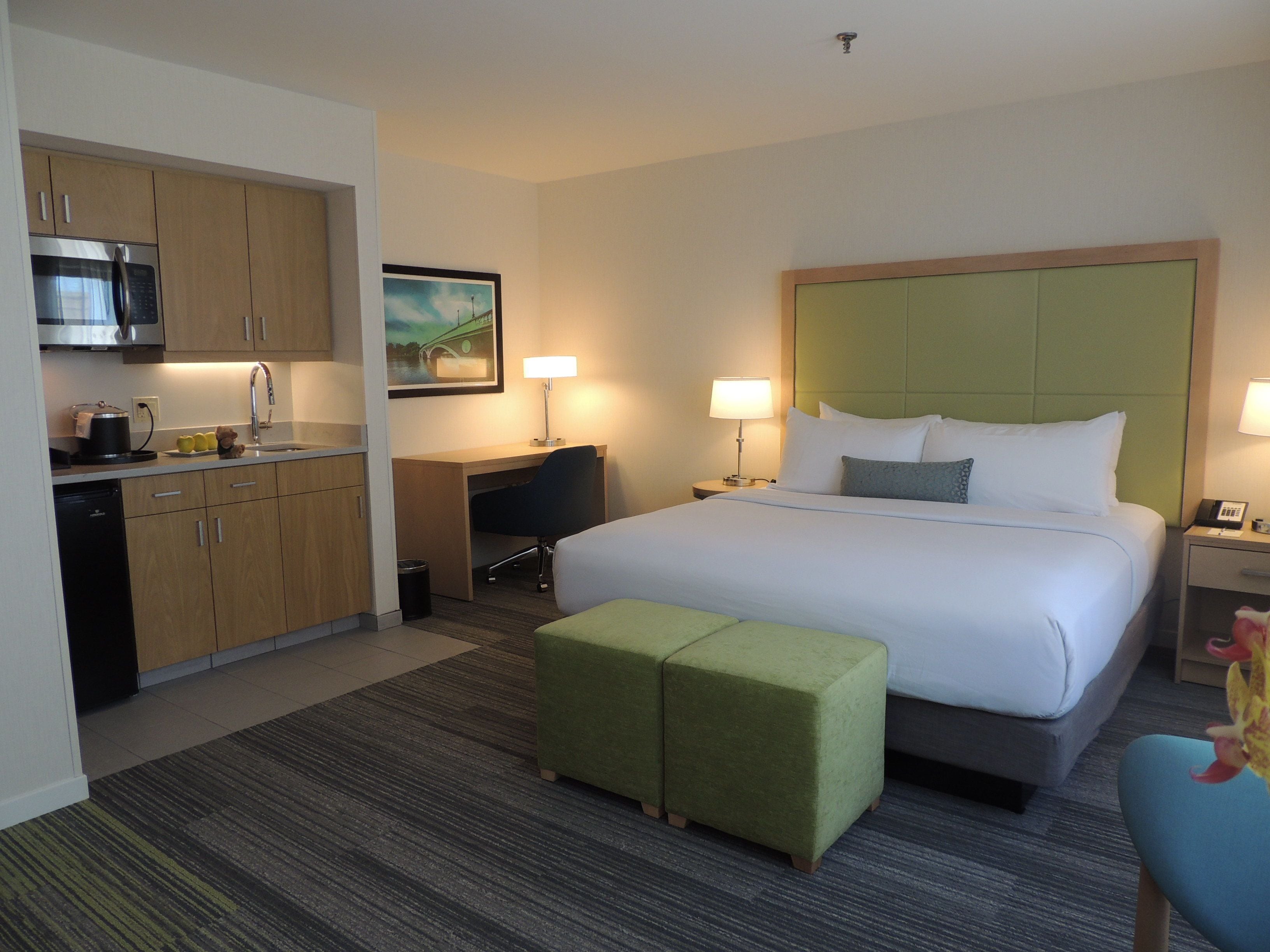 A hotel room with a bed, a desk, a kitchenette, and a green ottoman.