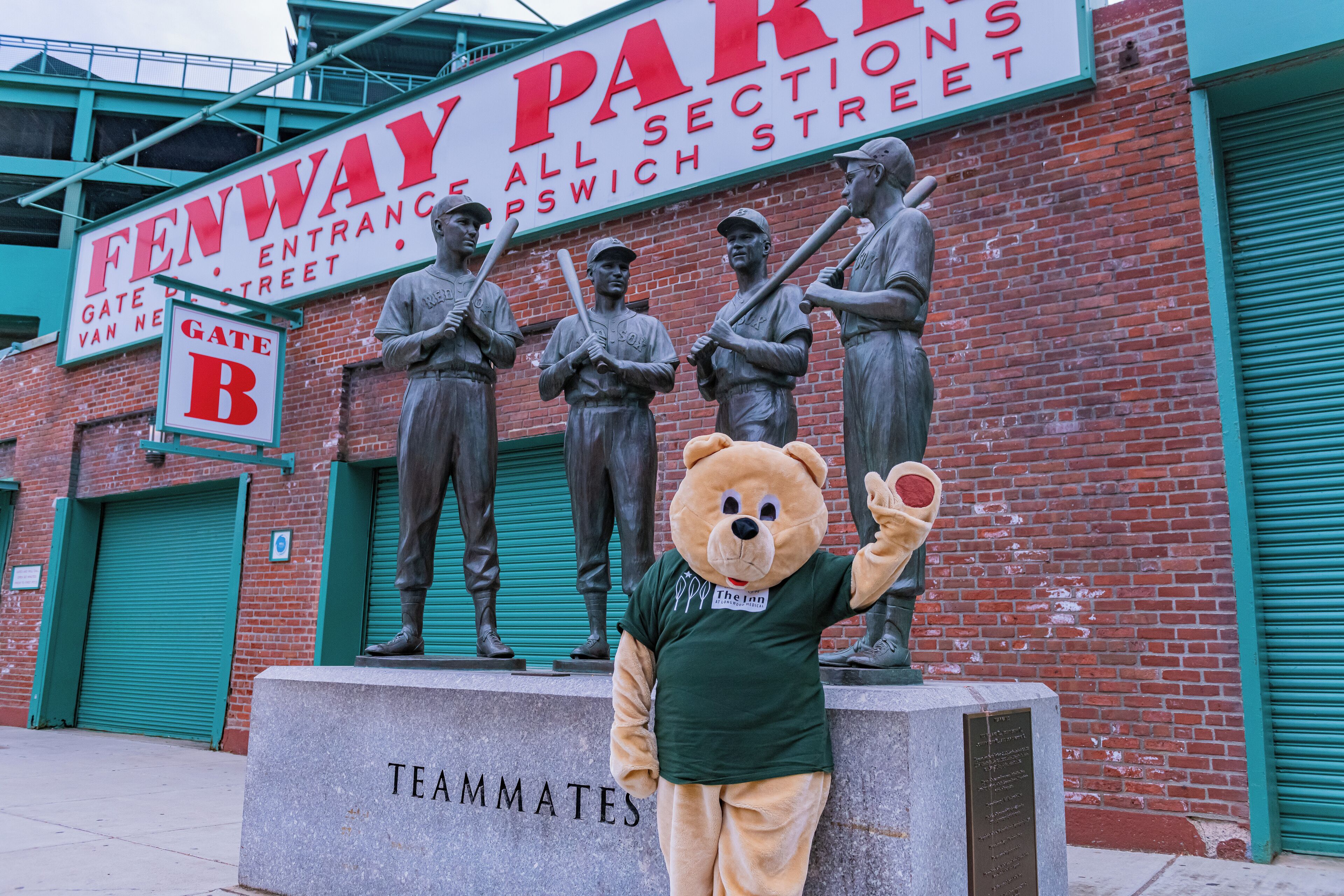 A mascot in front of a building with a sign reading "Fenway Park" and a "Gate B" sign.
