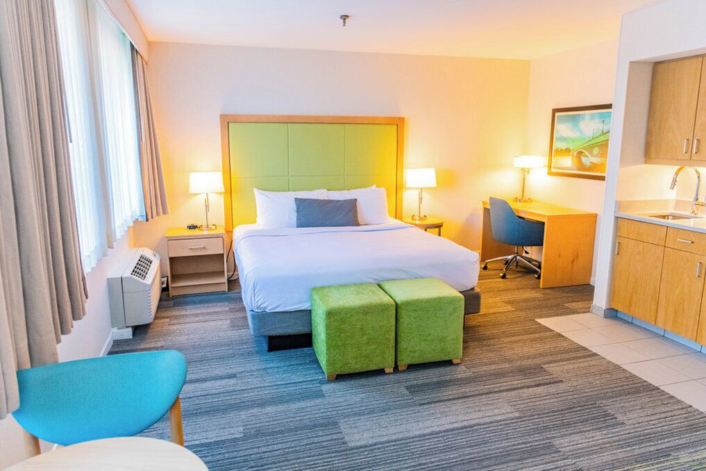A hotel room with a bed, two bedside tables, a desk, a chair, a kitchenette, and a window with curtains.