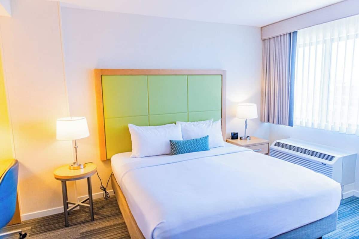 A hotel room with a large bed, two bedside tables with lamps, a window with curtains, and an air conditioning unit.