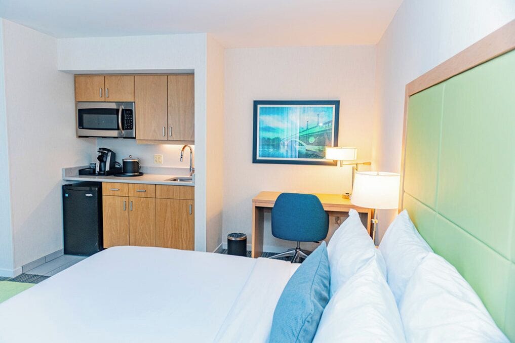 A hotel room with a bed, a desk with a chair, a microwave, a sink, and a trash can.
