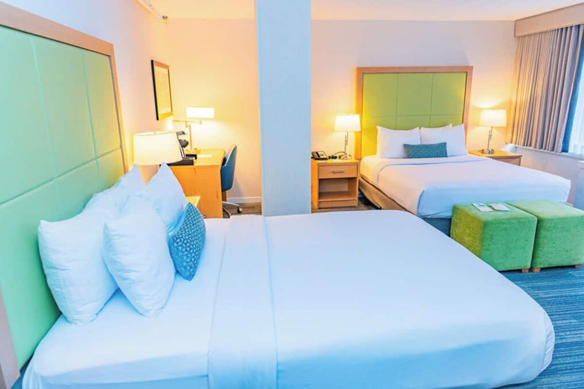 A hotel room with two beds, a desk, a chair, a nightstand, and a green ottoman.