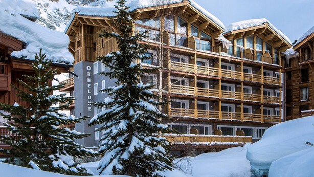 A multi-story wooden building with balconies, surrounded by snow-covered trees and mountains in the background. - Hotel Ormelune (Val-d'Isere)