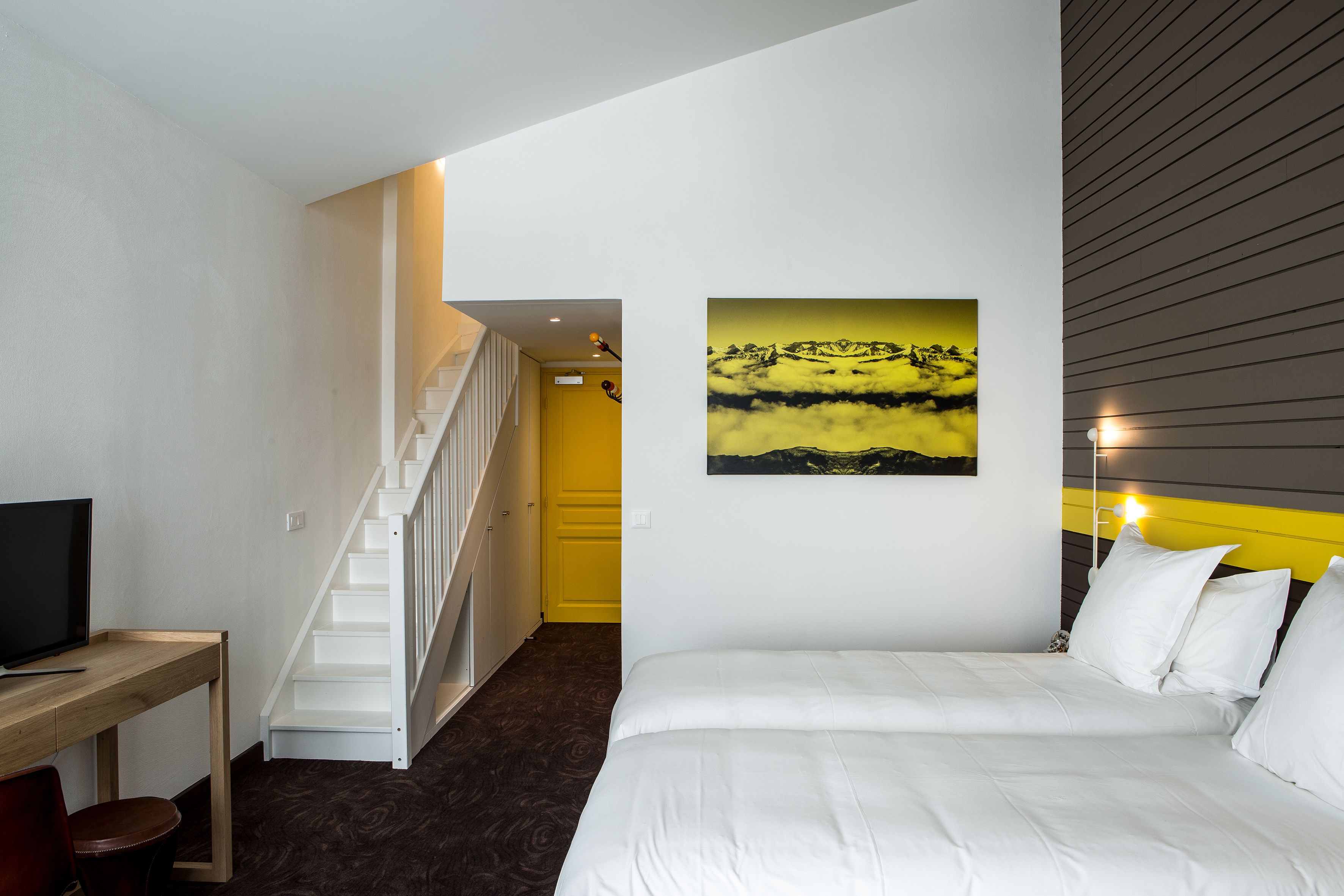 A bedroom with a bed, a desk, a television, and a staircase leading to a yellow room.