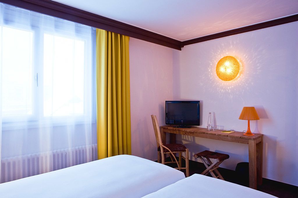 A hotel room with two beds, a desk with a television, a chair, a lamp, and a wall-mounted light fixture.