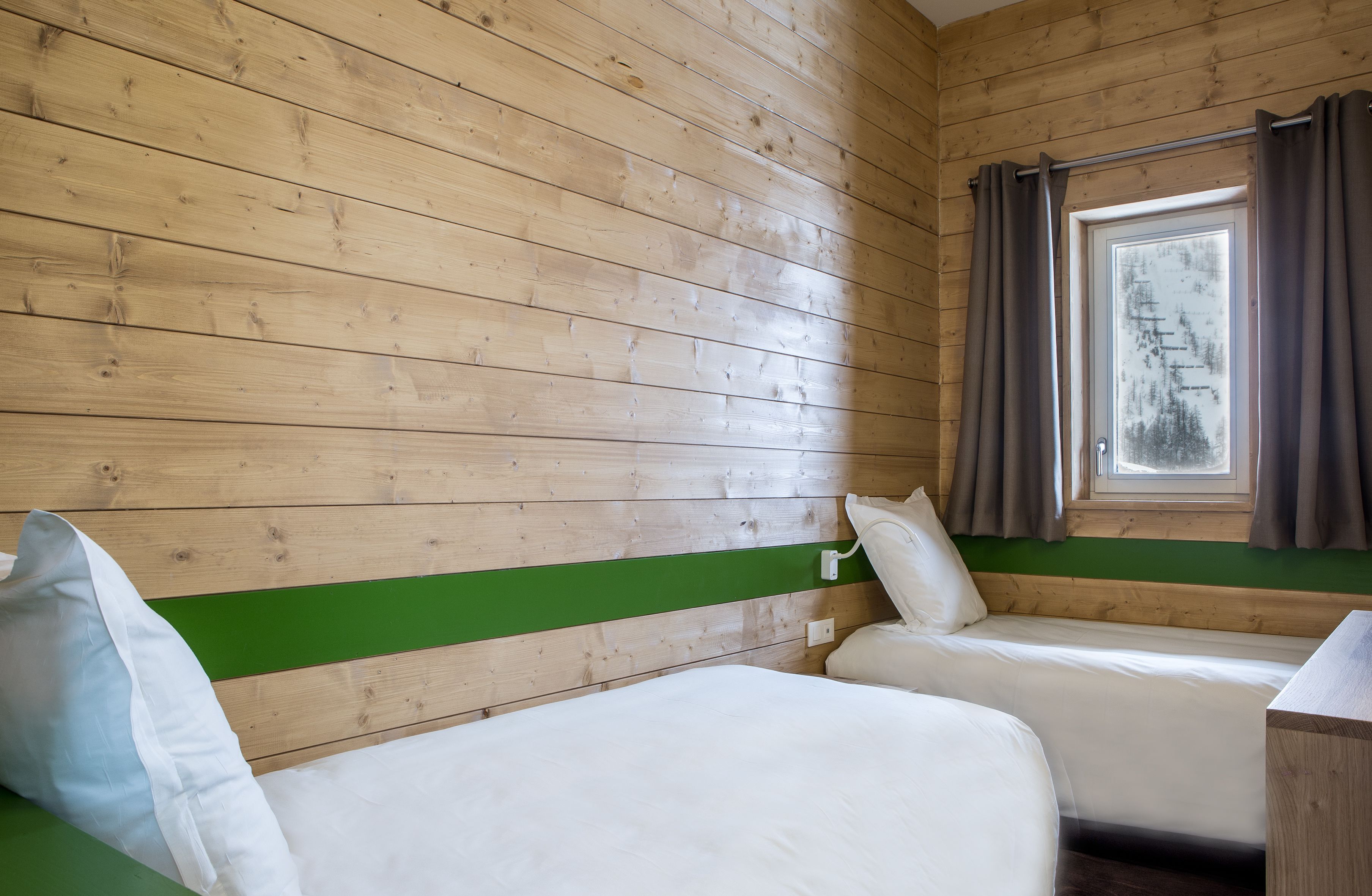 A cabin room with wooden walls, a green accent wall, two beds, a window with curtains, and a view of trees.