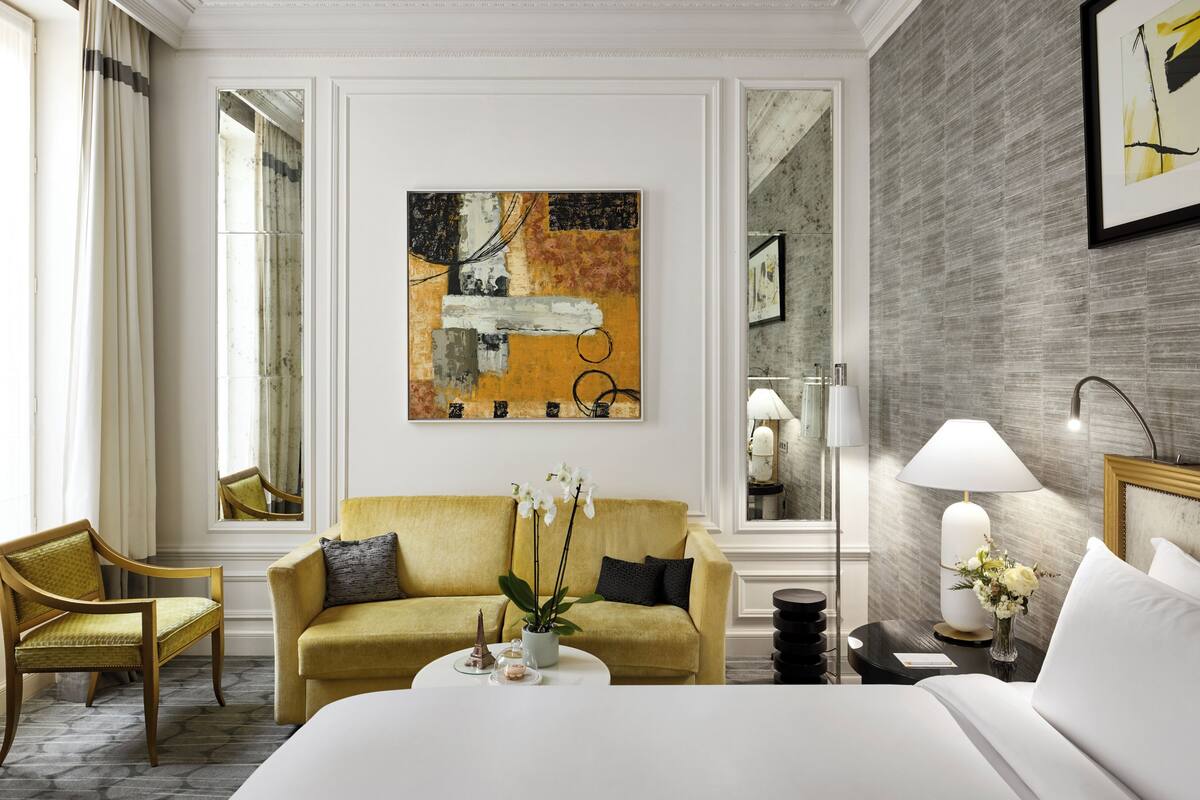 A hotel room with a yellow sofa, a bed, a coffee table, a lamp, and a painting on the wall.