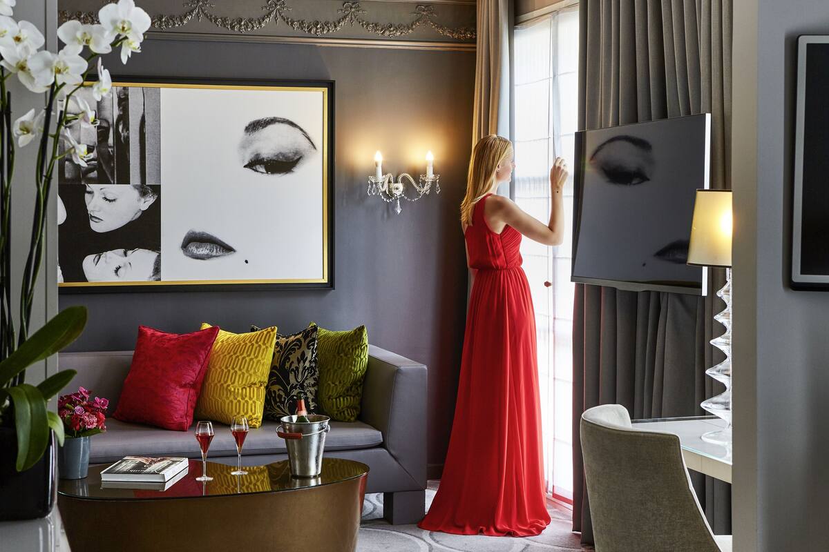 A modern hotel room with a grey sofa, red and yellow cushions, a glass coffee table with a book and two glasses, and a large mirror reflecting an eye.