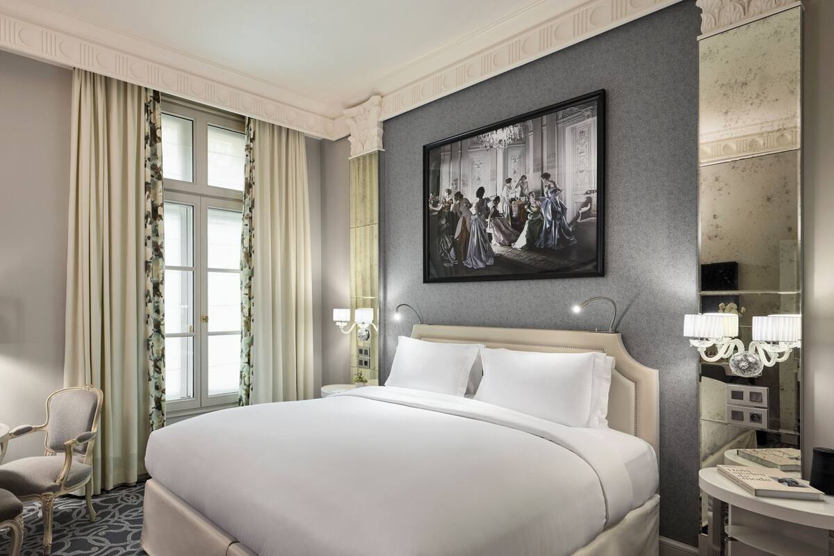 A hotel room with a large bed, a desk with a chair, a round table with a chair, a large mirror, and a painting on the wall.