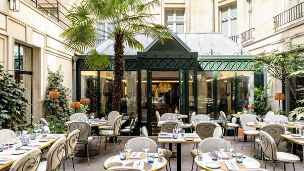 Un área de comedor al aire libre con mesas y sillas, una palmera y un edificio en el fondo.