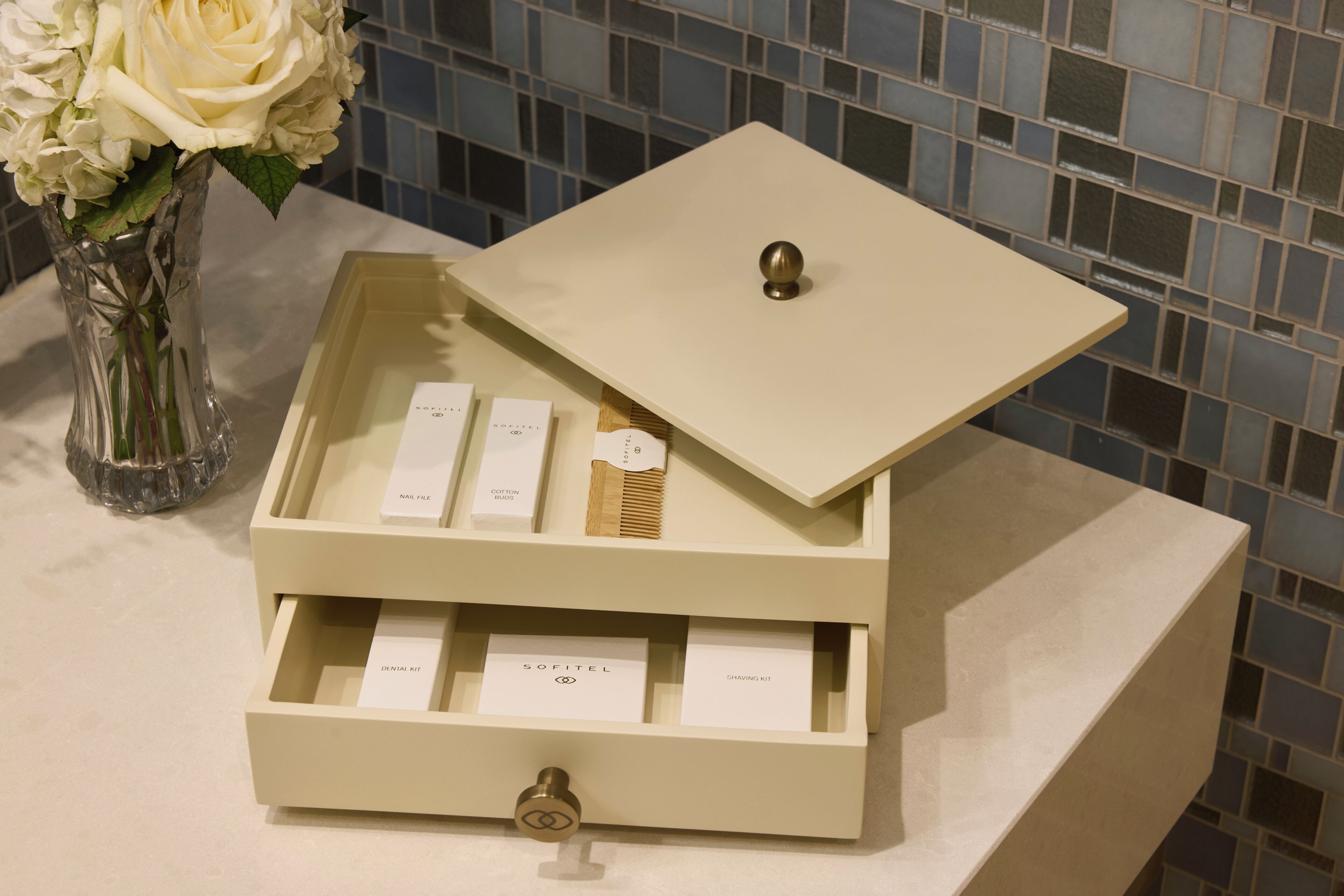 A bathroom vanity with a white marble countertop, a clear vase with white flowers, and a storage box with compartments labeled 'Nail File,' 'Softenail Kit,' and 'Shaving Kit.'