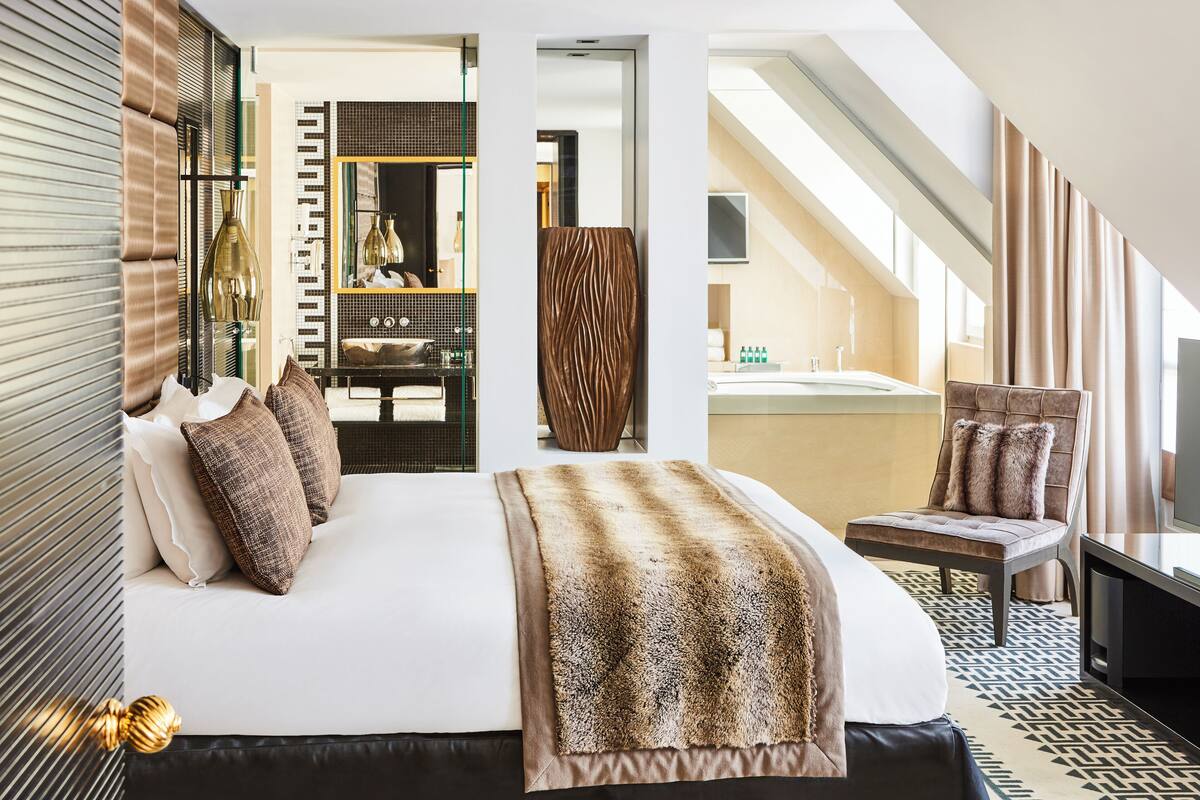 A modern hotel room with a large bed, a seating area with a chair, a television, and a bathroom with a bathtub and shower.