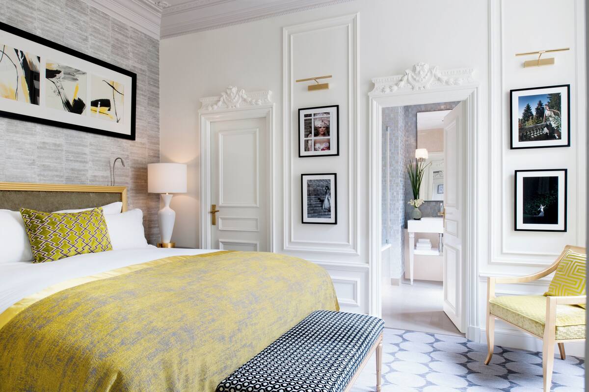 A hotel room with a large bed, a bench, a chair, a desk, and a bathroom visible through an open door.