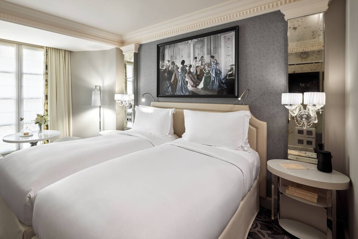 A hotel room with two beds, a sitting area with a chair and table, a large mirror, and a painting on the wall.