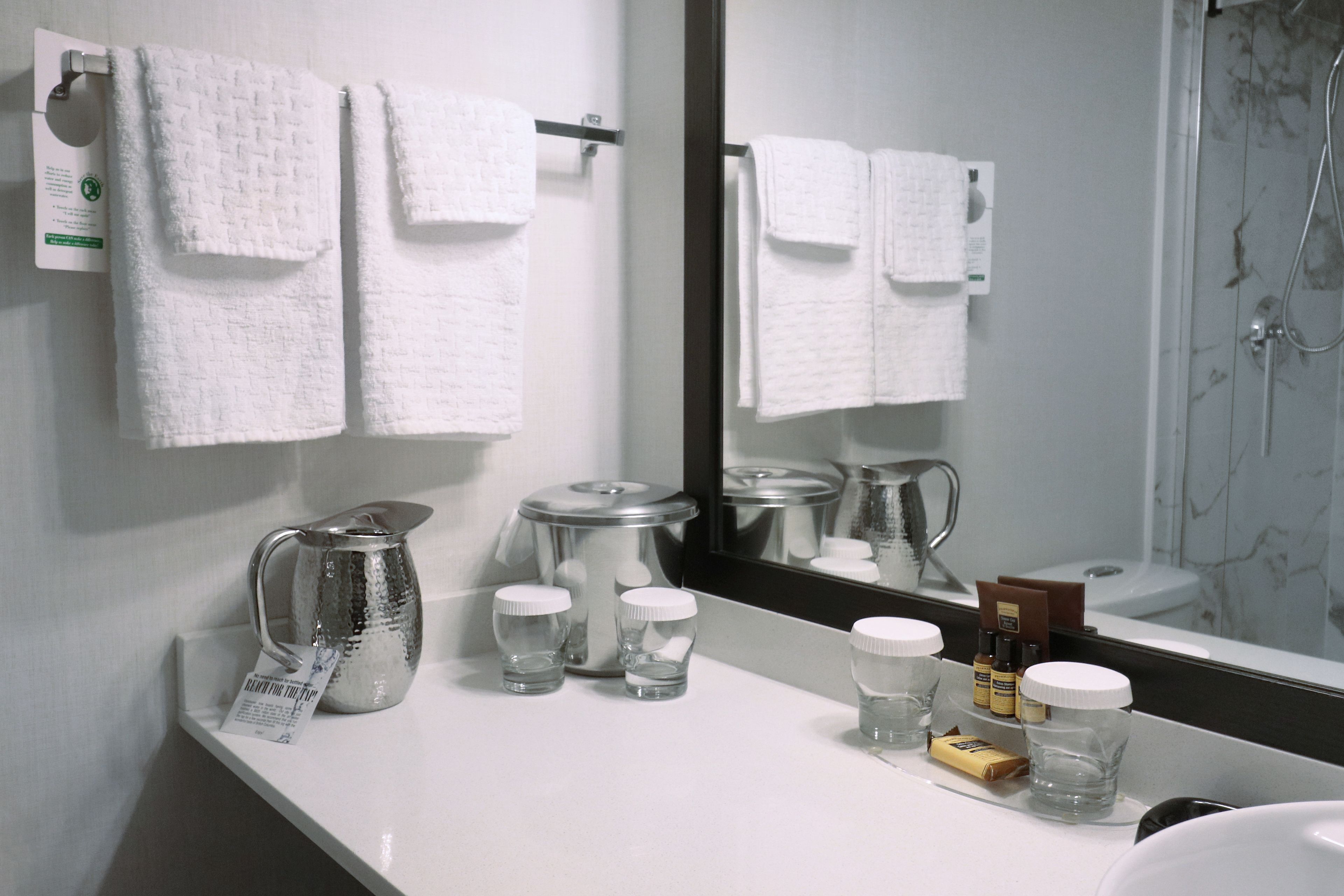 A bathroom with white towels hanging on a rack, a large mirror, a shower area, and a countertop with a kettle, shot glasses, and toiletries.