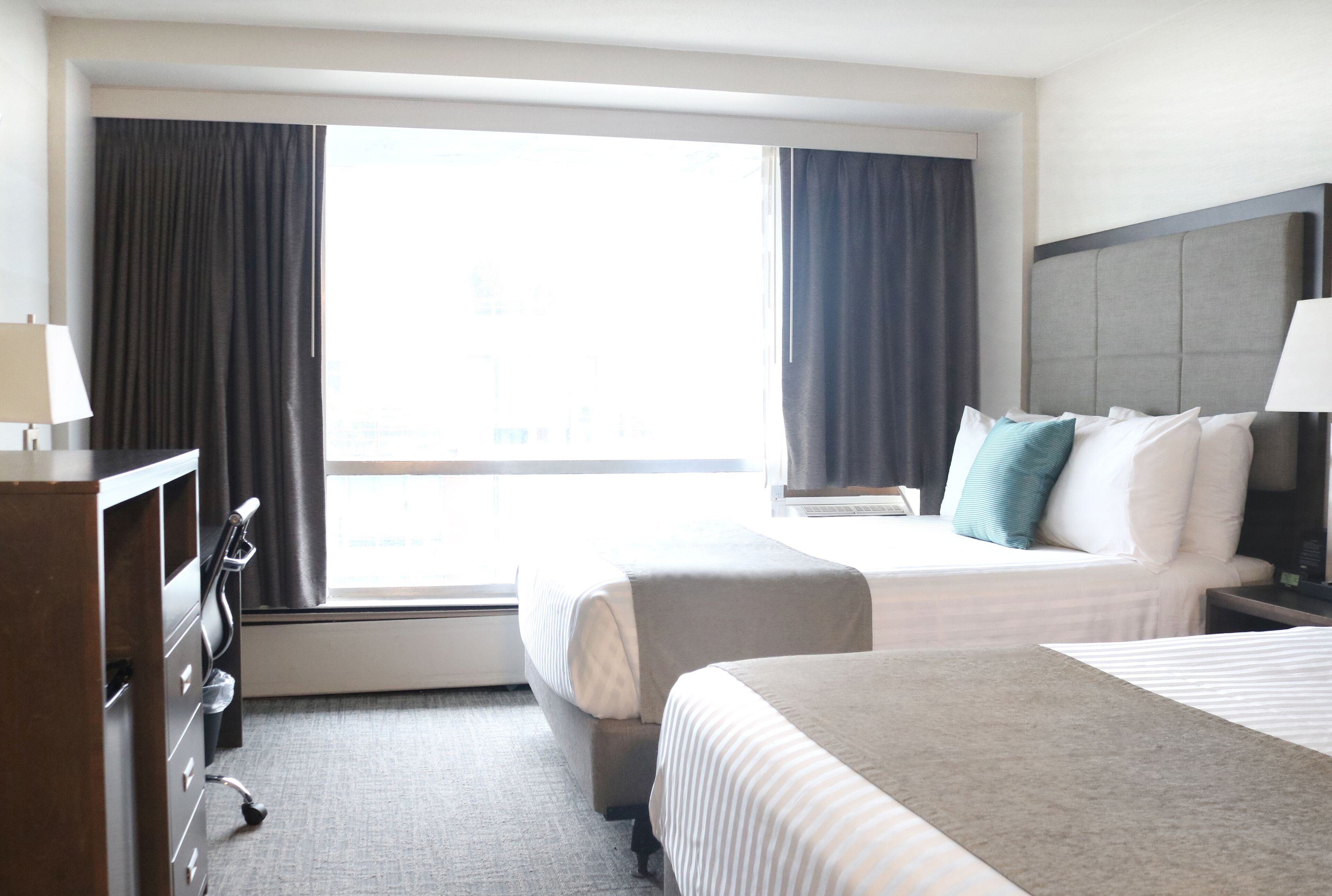 A hotel room with two beds, a dresser, a window with curtains, and a view of the city.
