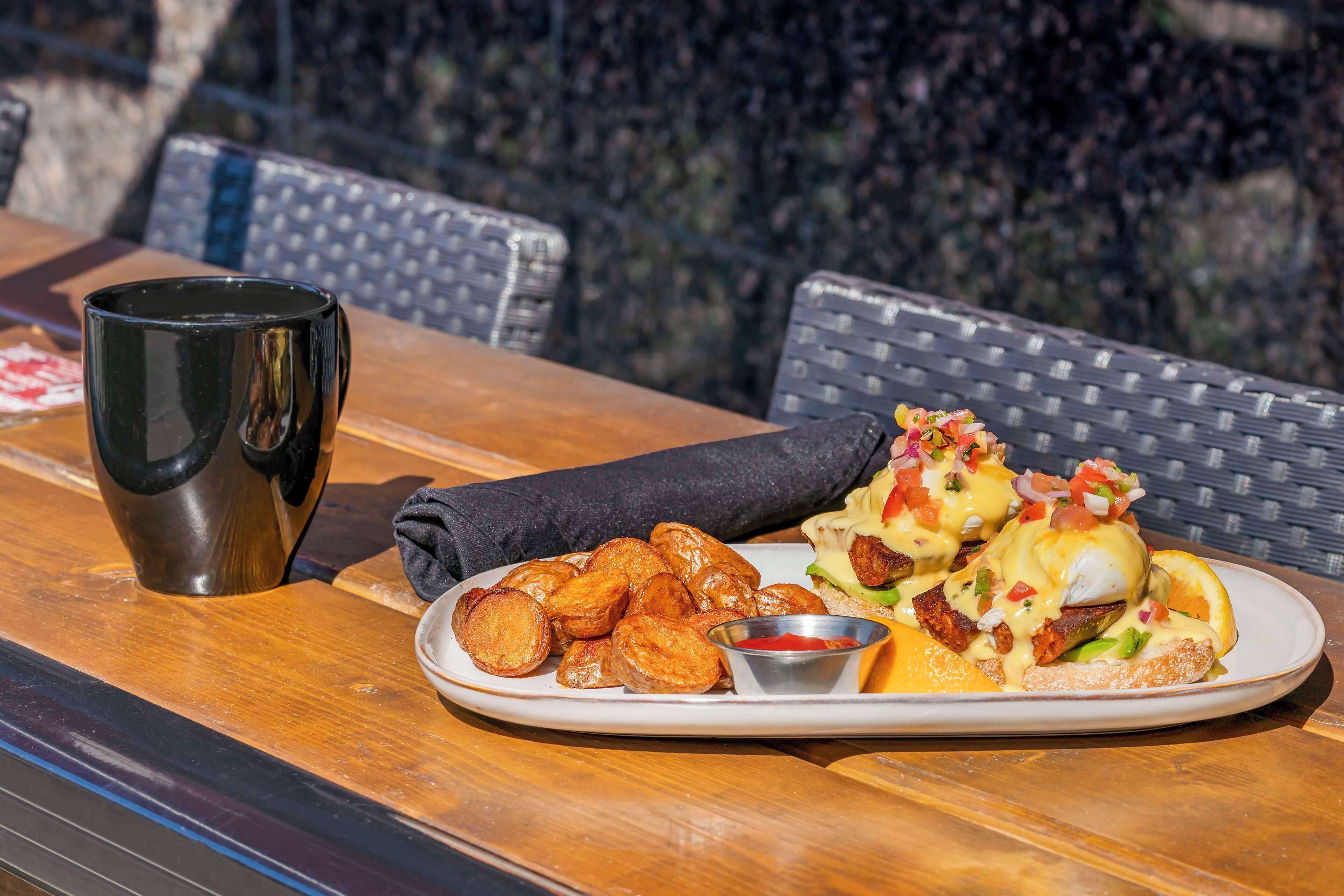 Un plato de comida que incluye huevos Benedictinos, una taza pequeña y una servilleta sobre una mesa de madera.