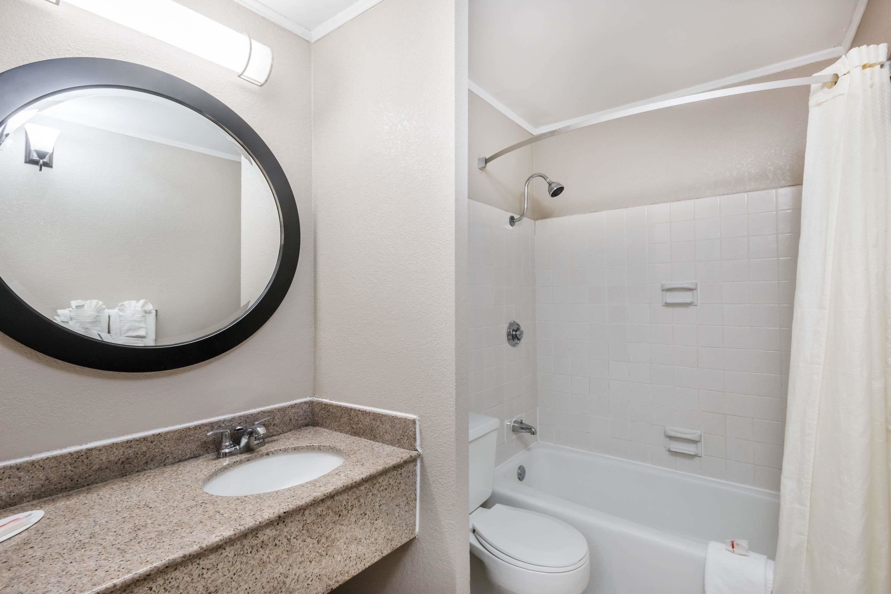 Un baño con lavamanos de encimera de granito, un espejo redondo, inodoro blanco y bañera con cabezal de ducha.