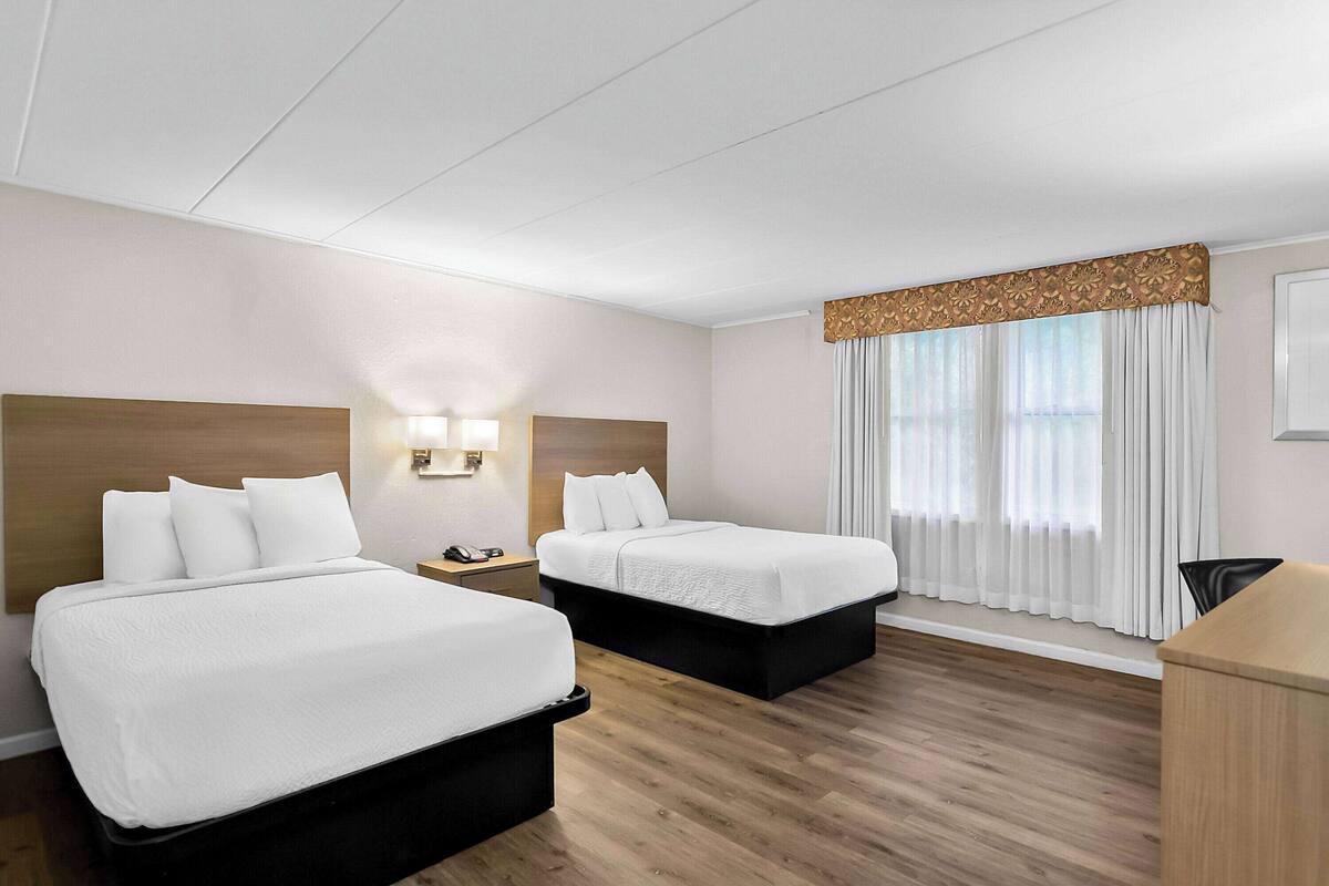 Une chambre d’hôtel avec deux lits, une tête de lit en bois, une table de chevet avec un téléphone, un bureau et une fenêtre avec des rideaux.