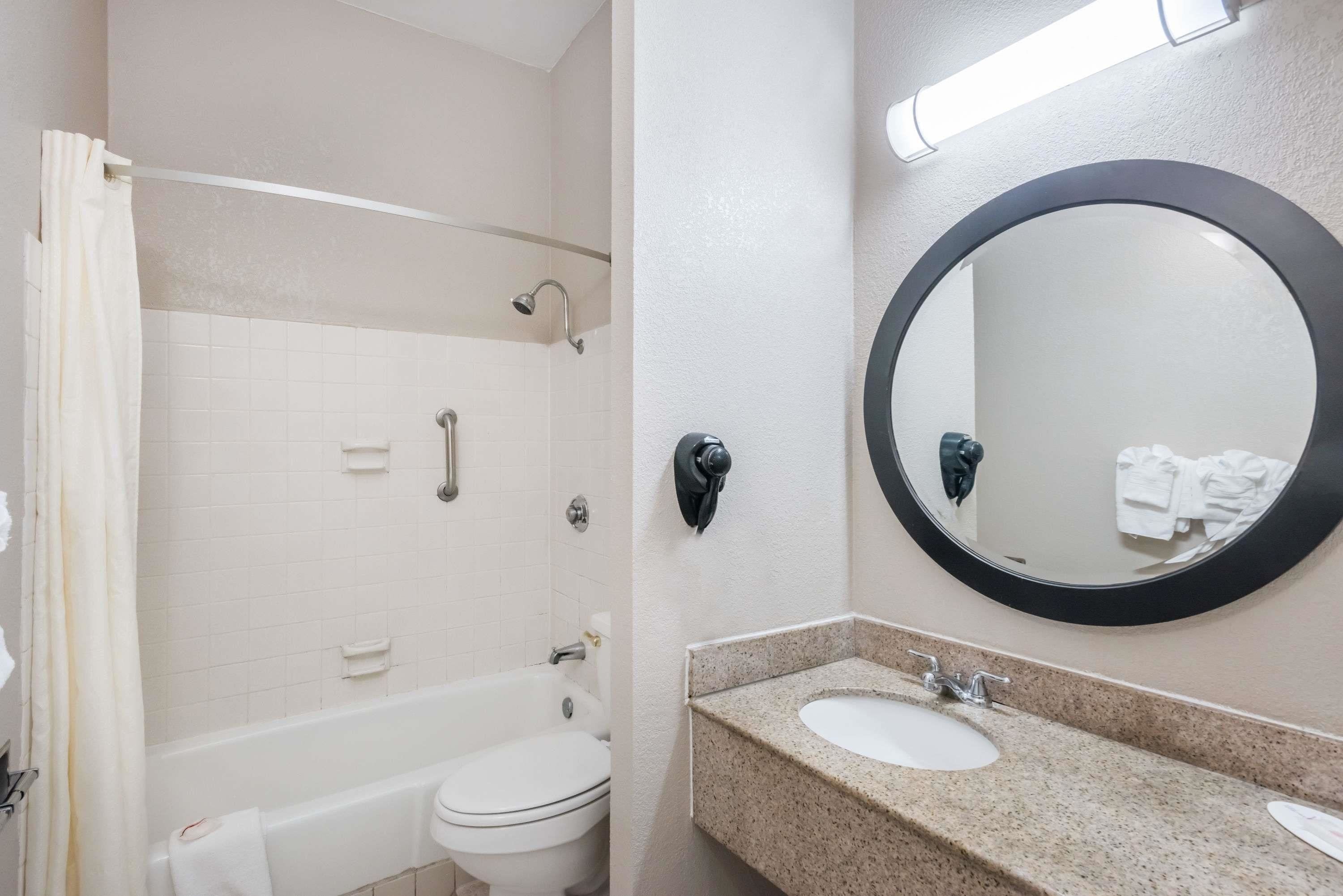 A bathroom with a white bathtub, a white toilet, a sink with a granite countertop, and a round mirror with a black frame.