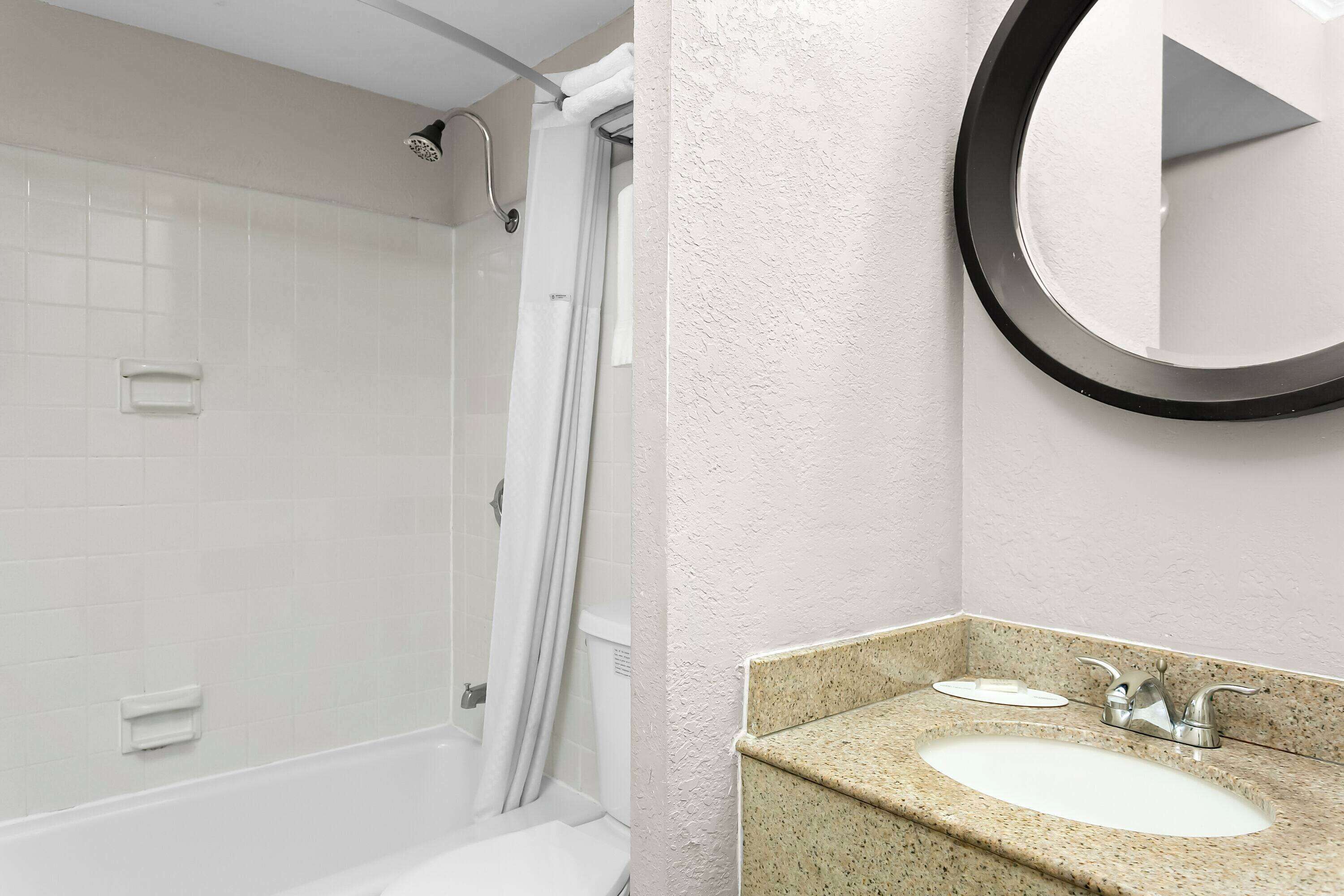 A bathroom with a shower, a sink with a granite countertop, and a round mirror.