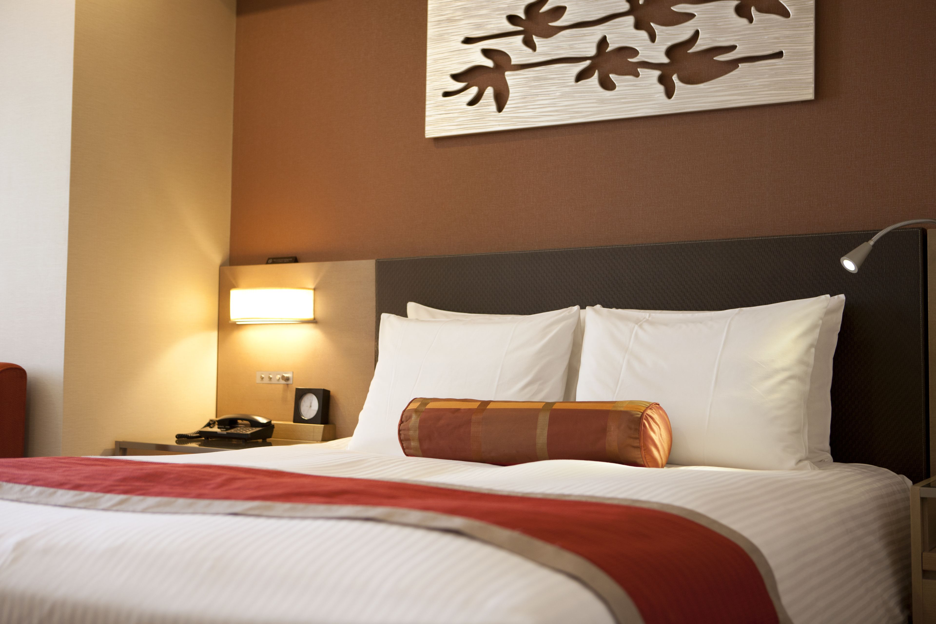 Una habitación de hotel con una cama bien tendida, una lámpara de noche, un reloj y una obra de arte en la pared.