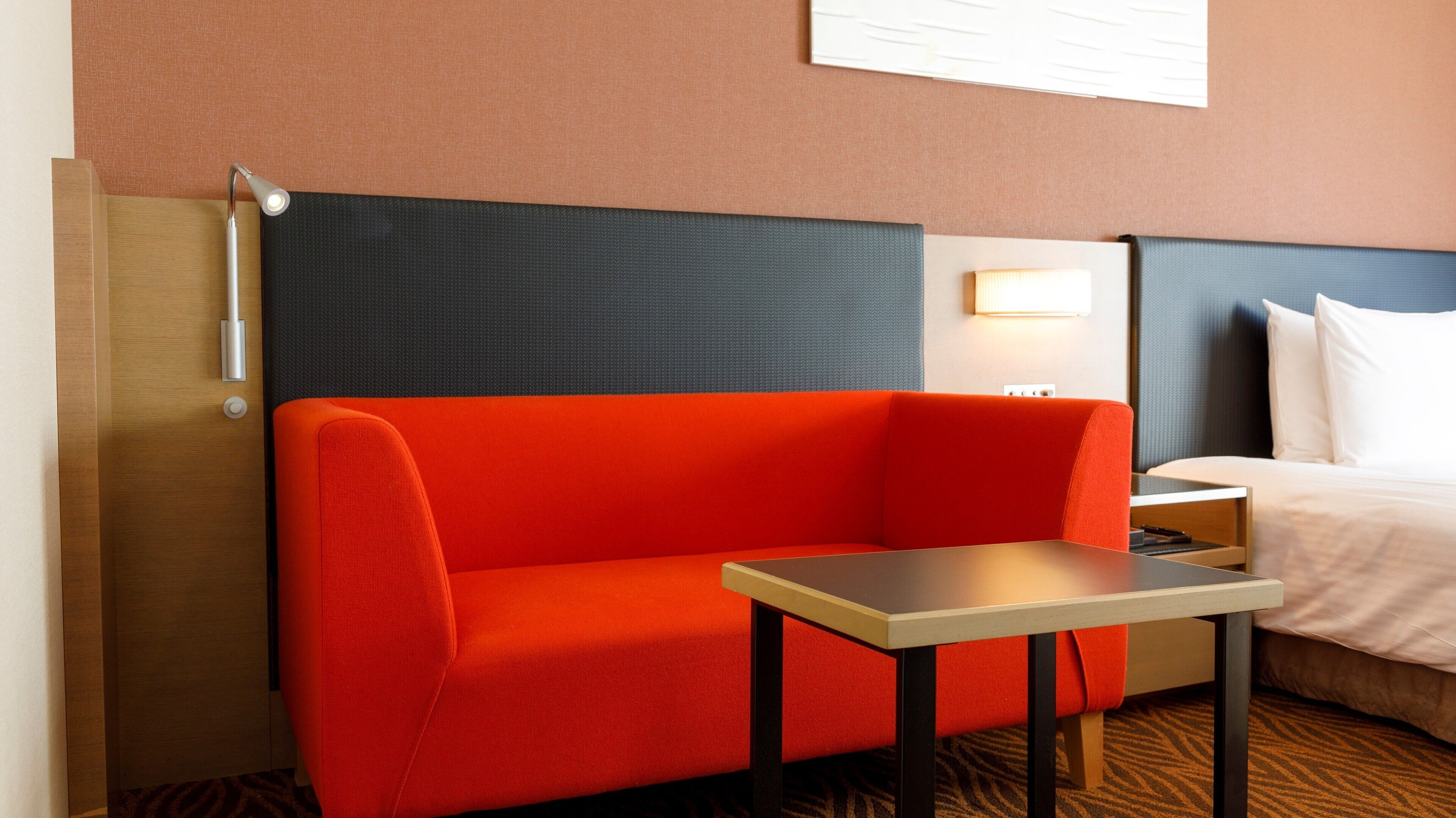 A hotel room with a red sofa, a small table, a bed with white linens, and a nightstand.