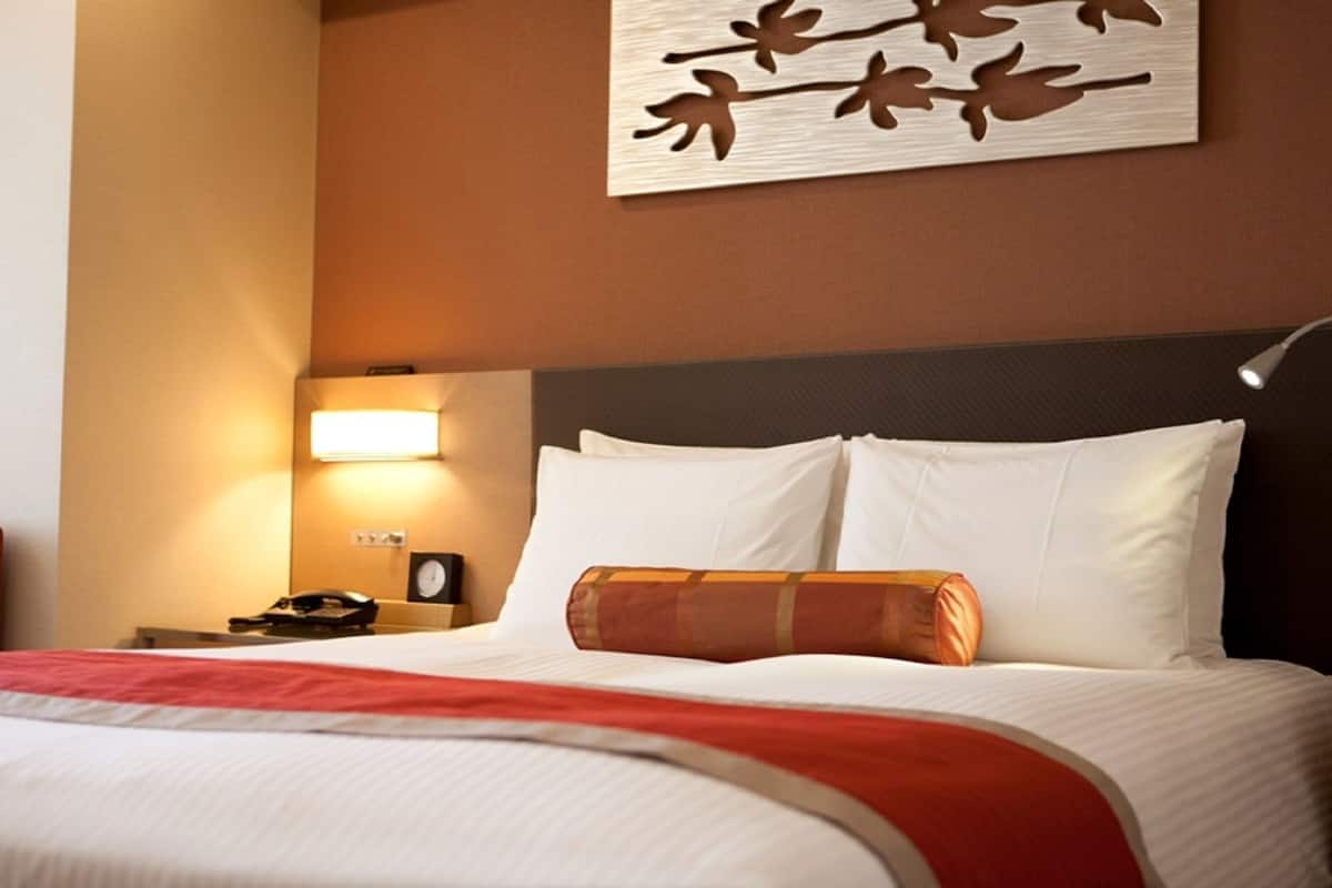 Quarto de hotel com uma cama, mesa de cabeceira com abajur, relógio e uma obra de arte na parede.