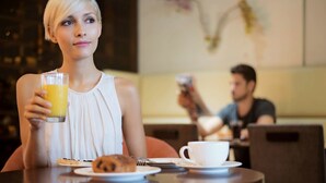 A person holding a glass of orange juice at a table with a croissant and a cup, in a dining area with another person in the background. - Novotel Montreal Centre (Montreal)