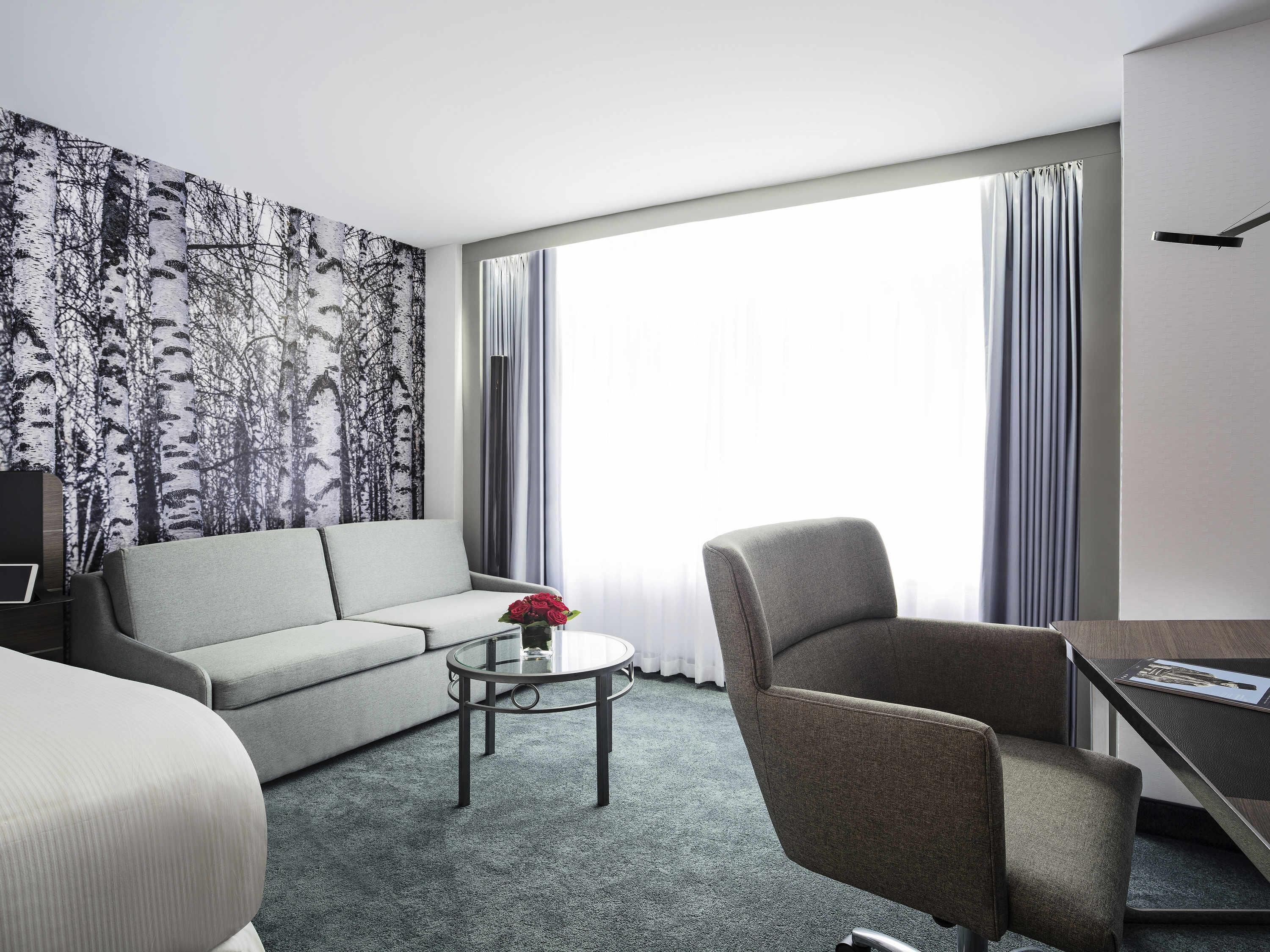 A hotel room with a sofa, armchair, small round table, and a large window with curtains.