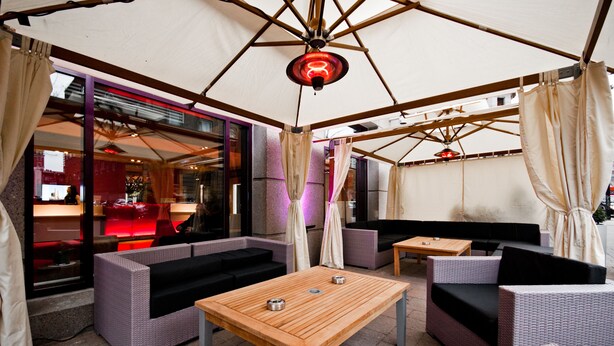 An outdoor seating area with wicker furniture, a wooden table, and a red neon light. - Novotel Montreal Centre (Montreal)